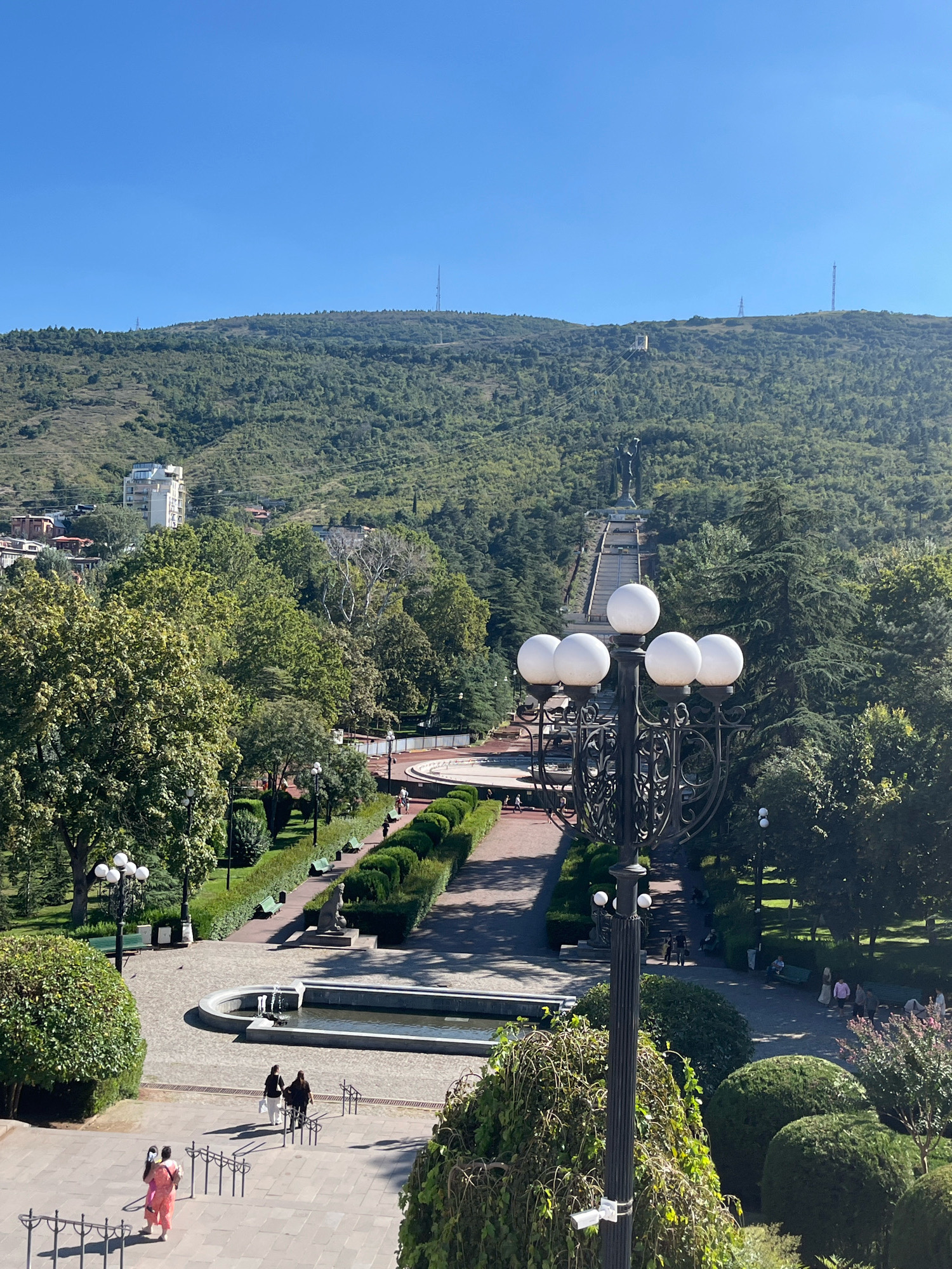 The image depicts a scenic view of a park area in Tbilisi, Georgia, looking up towards a monumental statue on a hillside. In the foreground, a prominent lamppost with multiple lights stands tall. Beyond the lamppost, a pathway leads through a meticulously maintained park with neatly trimmed hedges, benches, and a fountain. Several individuals are seen walking along the paths and enjoying the park. In the middle ground, a wide staircase ascends the hillside, leading to a large statue at the top. The statue appears to be of a figure standing with arms outstretched or holding an object. The hillside is densely covered with trees, and in the distance, buildings and communication towers are visible on the slopes. The sky is clear and blue, indicating a sunny day. The overall scene conveys a sense of public space and natural beauty within an urban environment.