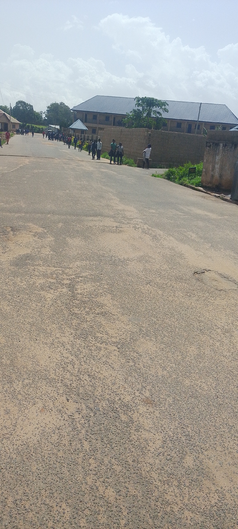 This media file depicts an outdoor scene in Bukuru, Nigeria, under a bright, cloudy sky, suggesting it is daytime. The foreground is dominated by an asphalt road with a textured, somewhat worn surface, reflecting the arid or dusty environment. In the mid-ground, a group of people, predominantly men and young adults, are walking along the side of the road. They appear to be in motion, possibly heading to or from a destination. Many are dressed in casual clothing, and some carry bags.
Behind the people, a long, multi-story building with a dark, sloped roof is visible. The building's facade is a muted brown or beige, and it appears to be constructed with brick or concrete. A low wall, possibly made of rough stone or concrete blocks, runs along the front of the building. Adjacent to this wall and the road, there is sparse greenery, with some bushes and trees adding touches of color to the scene.
To the right of the frame, a section of another wall is visible, made of rough, lighter-colored stone, which appears older or more weathered than the wall in front of the building. The overall impression is of a developing area or a community setting, possibly a school or residential compound, with people going about their daily activities on a warm day. No visible text is present in the media file.