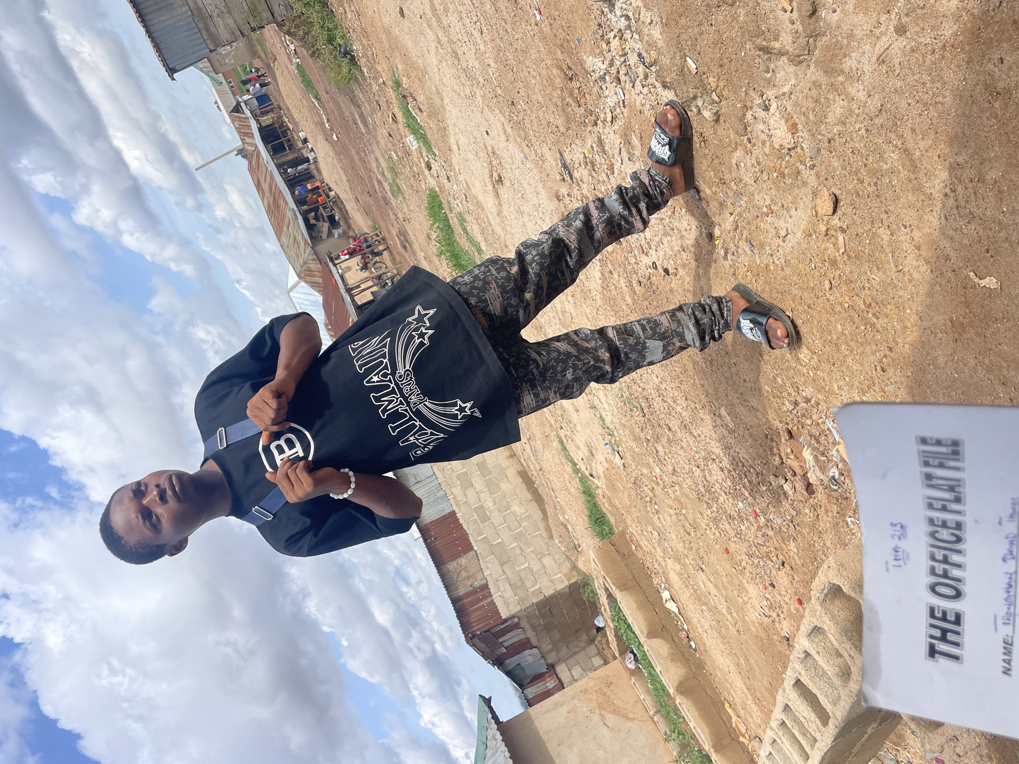 A young man stands in a dusty, outdoor area in Durumi, Nigeria. He is wearing a black Balmain t-shirt, patterned pants, and sandals. The background includes simple structures with corrugated metal roofs, suggesting a less affluent area. A document labeled "THE OFFICE FLAT FILE" is partially visible in the foreground. The environment appears to be a street or open space with a dirt surface and scattered debris. The sky is visible with patchy clouds.