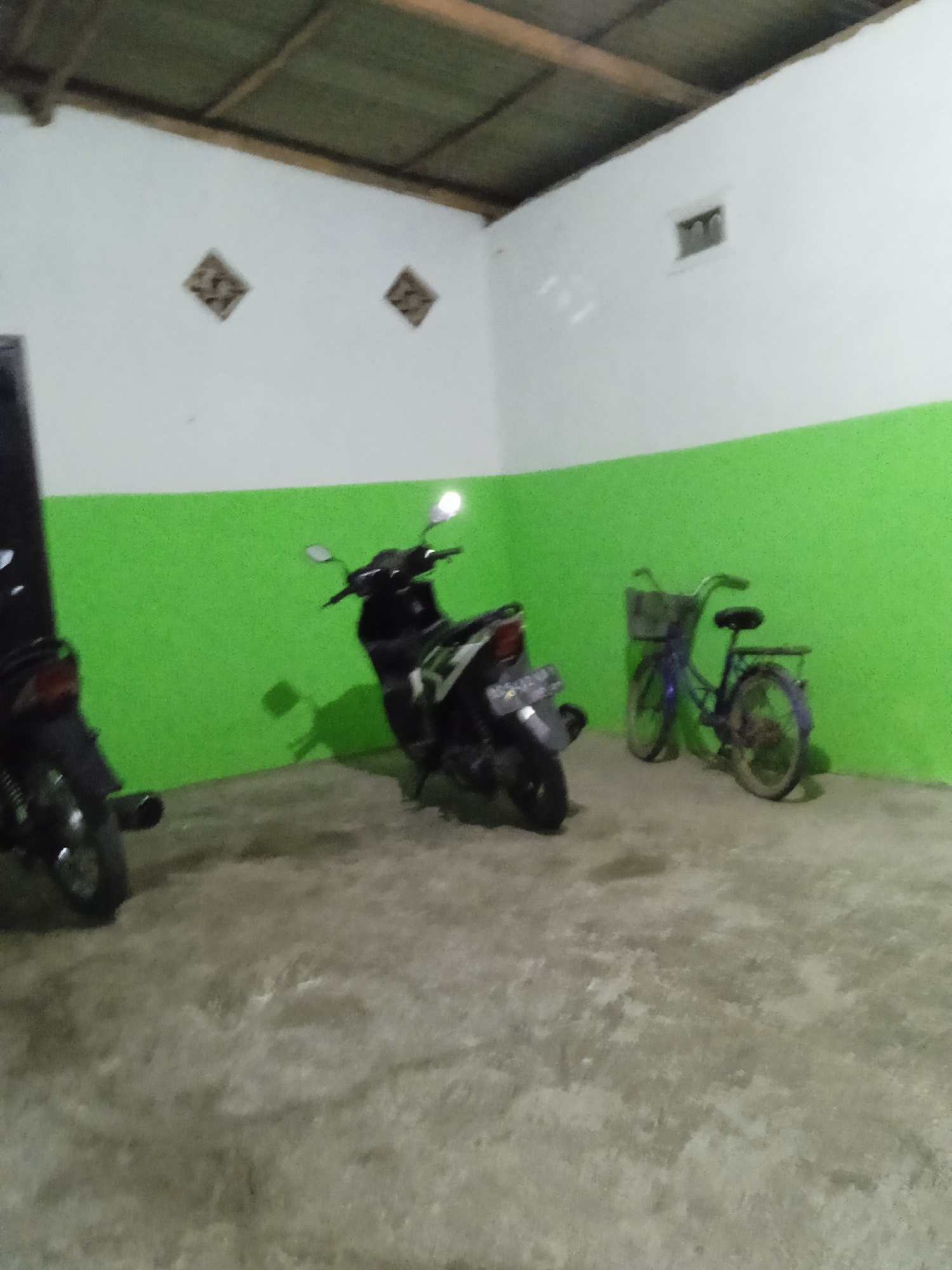 The image shows a covered parking area with two motorcycles and a bicycle parked on a concrete floor. The walls are painted white and green, and a corrugated metal roof is visible at the top. Two diamond-shaped decorative elements are present on the upper part of the white wall on the left, and a small rectangular vent is visible on the upper part of the white wall on the right. The lighting is dim, suggesting it is evening or night. The scene appears to be located in Kelurahan, Indonesia, based on the provided context.