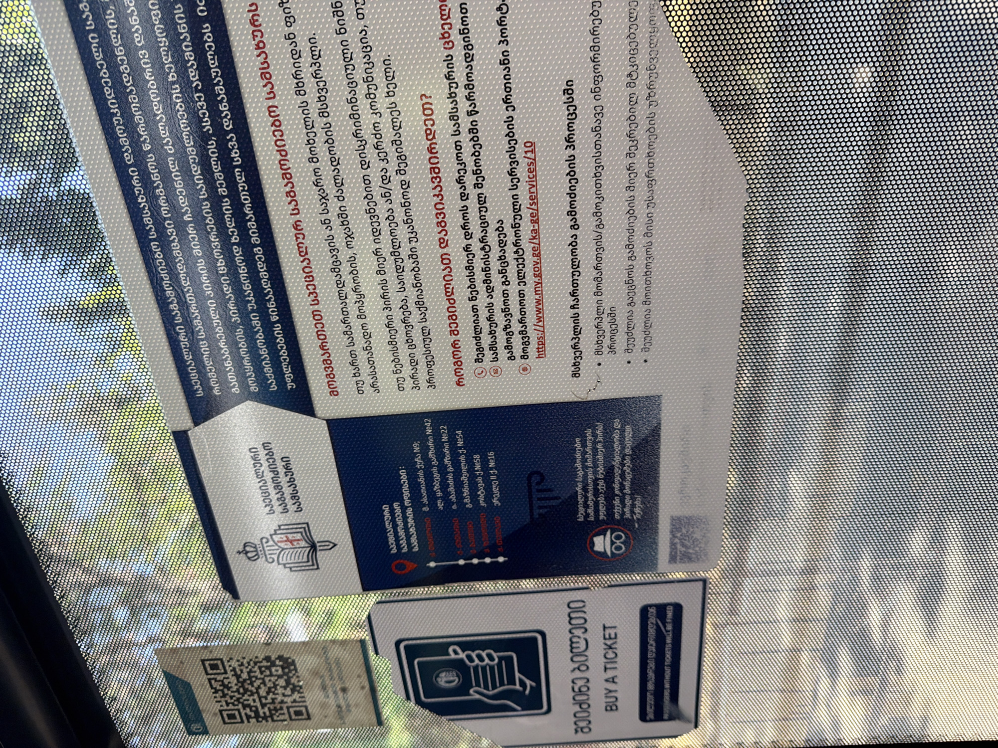 The image shows a close-up of a sign on a window, likely inside a public transportation vehicle in Tbilisi, Georgia. The sign is primarily in Georgian and contains text in both Georgian and English.
Key elements of the sign include:
* A section with Georgian text indicating "Information about public transport" and a QR code.
* A section with Georgian text titled "Travel regulations" and some numbered points detailing the regulations.
* A section with English text saying "BUY A TICKET" and "PASSENGERS WITHOUT TICKETS WILL BE FINED".
* Another section with Georgian text that appears to be contact information or directions to a location, possibly related to public transport. It includes a pin icon and the text "საინფორმაციო საკონტაქტო პუნქტი" (Information contact point) followed by a list of addresses or locations.
* A prominent image of a book with a sword piercing it, under a crown, which is the emblem of the Supreme Court of Georgia. This suggests a connection to a legal or governmental entity, possibly related to the enforcement of transport regulations or information dissemination.
* A graphic of a person in a hat with glasses, possibly representing a detective or inspector, next to another icon with two concentric circles and a line through them, likely indicating a restriction or prohibition.
The environment is characterized by the perforated texture of the window, through which a blurred outdoor scene of trees and sunlight is visible, suggesting the sign is placed on a window in a moving vehicle or a building with a screen-printed window. The overall impression is an informational poster related to public transport regulations and ticketing in Tbilisi.