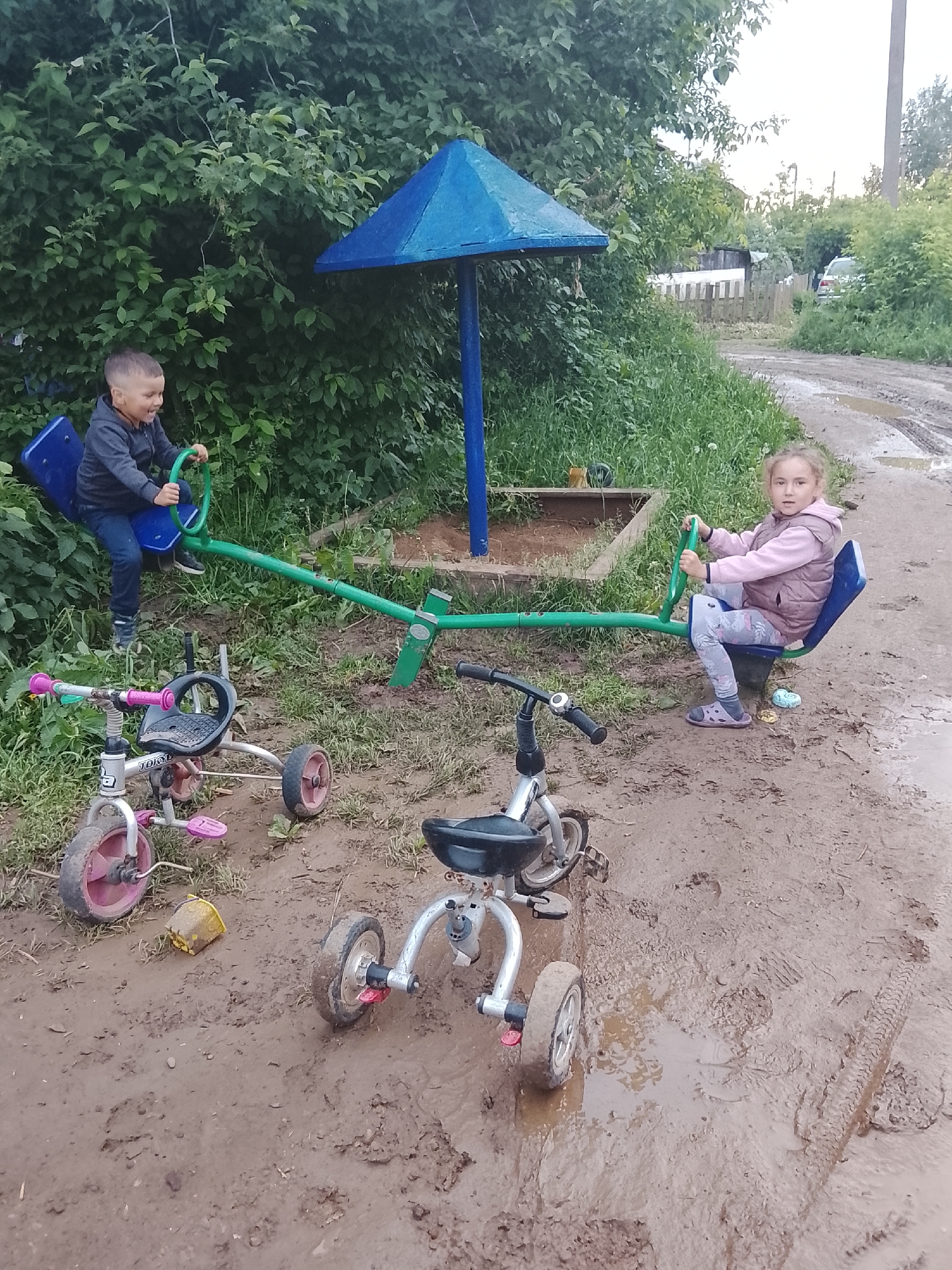 In the city of Kuyeda, Russia, two children are observed playing on a seesaw in an outdoor, muddy environment.
The scene features a young boy and a young girl. The boy, approximately 4-6 years old, is seated on the left end of a green seesaw, holding its green handle. He wears a dark grey hooded sweatshirt and dark blue jeans, with dark footwear. His expression indicates laughter or a smile, and his side of the seesaw is currently in a downward position. The girl, approximately 5-7 years old, is seated on the right end of the seesaw, also holding its green handle. She wears a light pink long-sleeved top, a light brown/pink puffer vest, grey patterned leggings, and dark purple sandals. Her end of the seesaw is in an upward position, and she is looking directly towards the camera with a neutral expression.
The seesaw is green metal with blue plastic seats and is anchored in a patch of dirt and sparse grass. Behind the seesaw's fulcrum, a blue conical structure on a blue pole stands over a small excavated area, possibly a sandpit. Dense green foliage forms a backdrop directly behind the seesaw.
In the foreground, two child-sized tricycles are visible on the muddy ground. The tricycle on the left features a silver frame with pink wheels and pedals, and a black seat; a yellow plastic cup lies overturned nearby. The tricycle on the right has a silver frame, black seat, and dark wheels. Both tricycles are upright and partially covered in mud.
The ground across the entire visible area is heavily saturated with mud, exhibiting numerous puddles, tire tracks (including a distinct tread mark on the far right), and footprints. The background on the right side reveals a dirt path extending into the distance, bordered by overgrown grass, a wooden fence, and scattered structures, including a partially visible light-colored car and a utility pole. The lighting suggests an overcast sky.