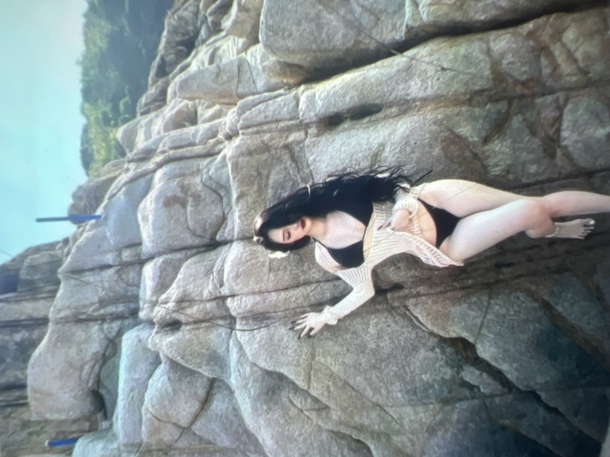 A young woman with long black hair, wearing a black bikini and a beige knit cover-up, poses against a large, textured rock formation. She is leaning with her left arm against the rock, her right arm extended downwards. The background consists of more rocks, some distant greenery, and a bright sky. The image appears to be taken outdoors, likely in a rocky, coastal, or mountainous environment. There is no information available to associate this image with the city of Kara-Dıykan, Kyrgyzstan.