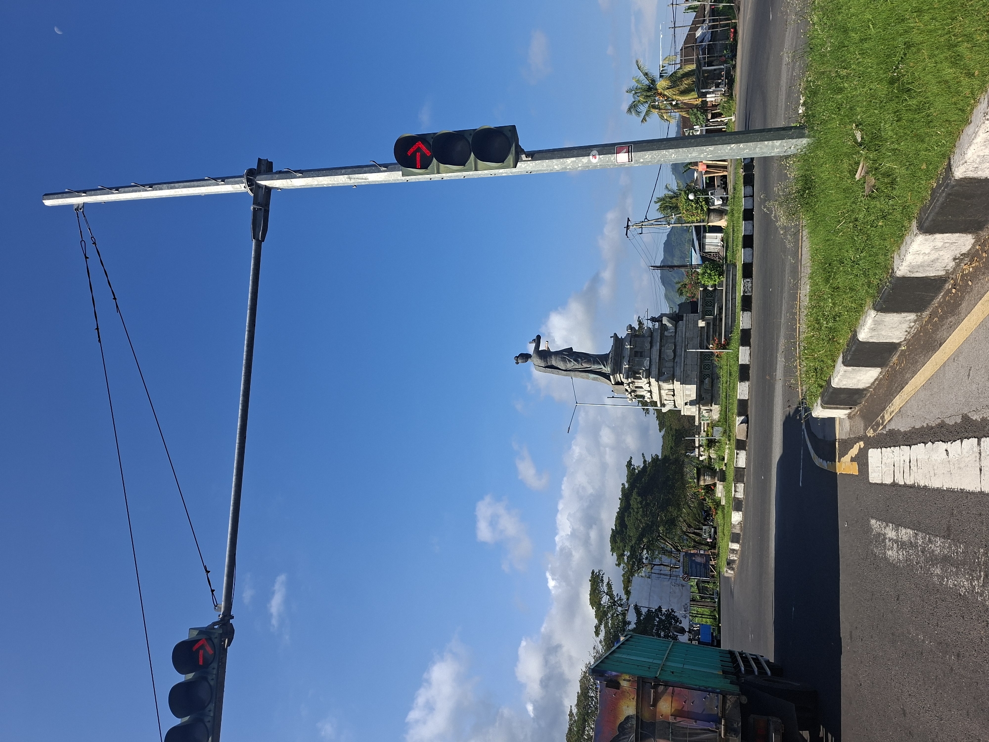 This image depicts a traffic intersection with a statue in the background. In the foreground, a traffic light with a red arrow signal is visible on a metal pole. A truck is partially visible on the left side of the image. The intersection appears to be a roundabout, with grassy medians and decorative borders. In the background, a tall, stone statue of a woman in traditional attire stands on a pedestal. Beyond the statue, buildings and lush green trees can be seen, with mountains in the distance. The sky is a clear blue with a few scattered clouds. This scene is located in the city of Dawan, Indonesia.