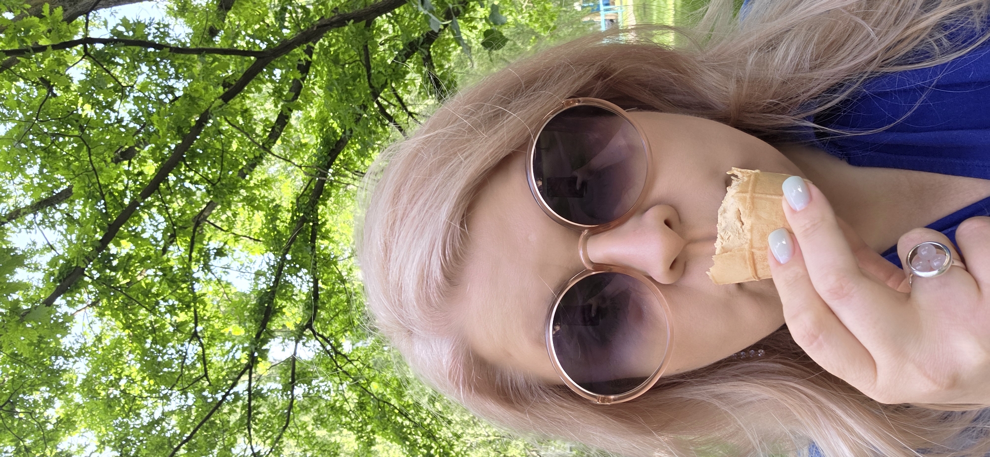 The original image is presented horizontally, rotated 90 degrees clockwise. It features a person, likely female, positioned prominently in the foreground against a backdrop of bright green foliage and tree branches.
The person has long, light blonde or platinum hair and fair skin. They are wearing large, round sunglasses with light brown or rose gold frames, which obscure most of their face. A blue top or dress is visible on their upper body. The person is holding a partially eaten waffle cone to their mouth, suggesting they are consuming a dessert like ice cream. Their visible fingers have light blue or white nail polish, and a silver-toned ring with a circular setting containing three small, light pink spherical stones is worn on their left ring finger. A reflection in the left sunglass lens shows a hand holding a smartphone, indicating the image is a selfie taken by the subject.
The background consists of dense, vibrant green leaves and tree branches, with patches of bright blue sky peeking through, indicating an outdoor setting such as a park or garden. The bright lighting suggests it is daytime, likely a sunny and clear day. The activity depicted is casual and relaxed, enjoying a treat in a natural environment. There is no visible text in the image.