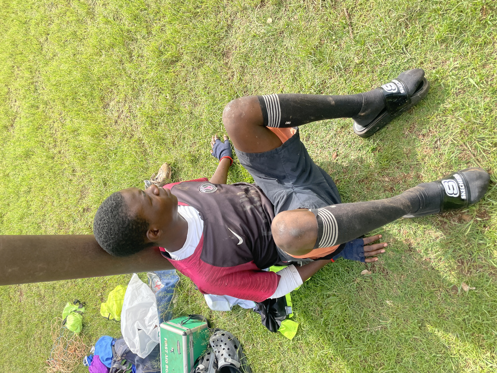 A young Black male is sitting on a grassy area, leaning against a brown, cylindrical pole, in what appears to be a park or sports field. He is wearing a short-sleeved, black and red soccer jersey with a white undershirt, dark shorts with orange lining, black socks with white stripes, and black slip-on sandals. He has on dark blue gloves on his left hand and a red wristband on his right. His left leg is bent at the knee, and his right leg is extended slightly. Several bags, a green first-aid box, and some ropes are scattered on the grass around him. The overall scene is well-lit, suggesting it is daytime. The presence of a first-aid box and athletic wear implies a potential sports-related activity or break.