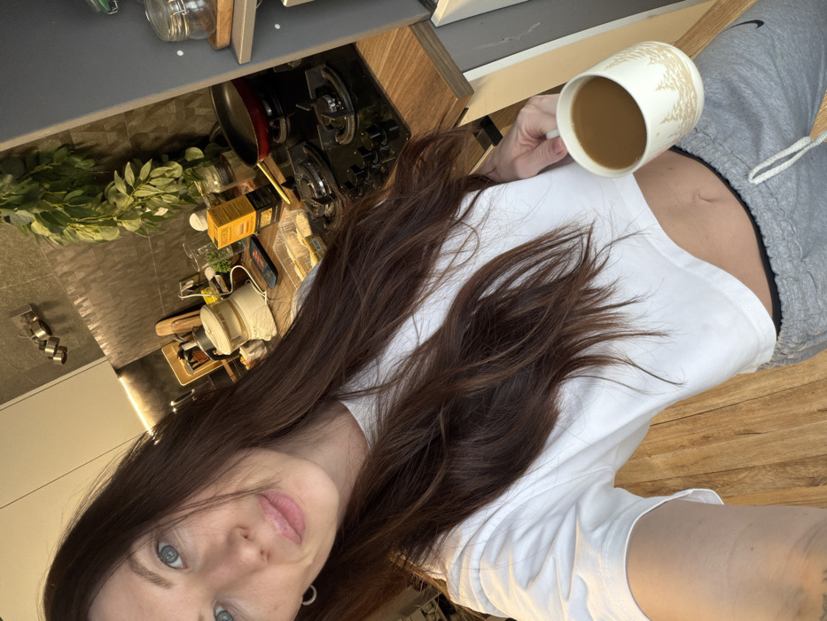 A female subject is depicted from the chest up, taking a self-portrait in a kitchen located in Perm, Russia. She possesses long, dark brown hair that falls over her left shoulder and chest, and blue eyes. Her lips are slightly parted in a faint smile. She wears a white, short-sleeved crop top that exposes her midriff, paired with grey sweatpants featuring a visible white drawstring. Her right hand holds a white ceramic mug adorned with a light brown tree pattern, containing a light brown liquid.
The kitchen background features a dark grey tiled or textured backsplash and a wood-grain countertop. Visible on the countertop are a white food steamer/processor, a black gas cooktop with a red frying pan, a yellow packaged item, and a clear container holding white contents. A green leafy plant is draped against the backsplash. Overhead white cabinetry and a wall-mounted metallic spice rack are also visible. The floor is light wood-grain.