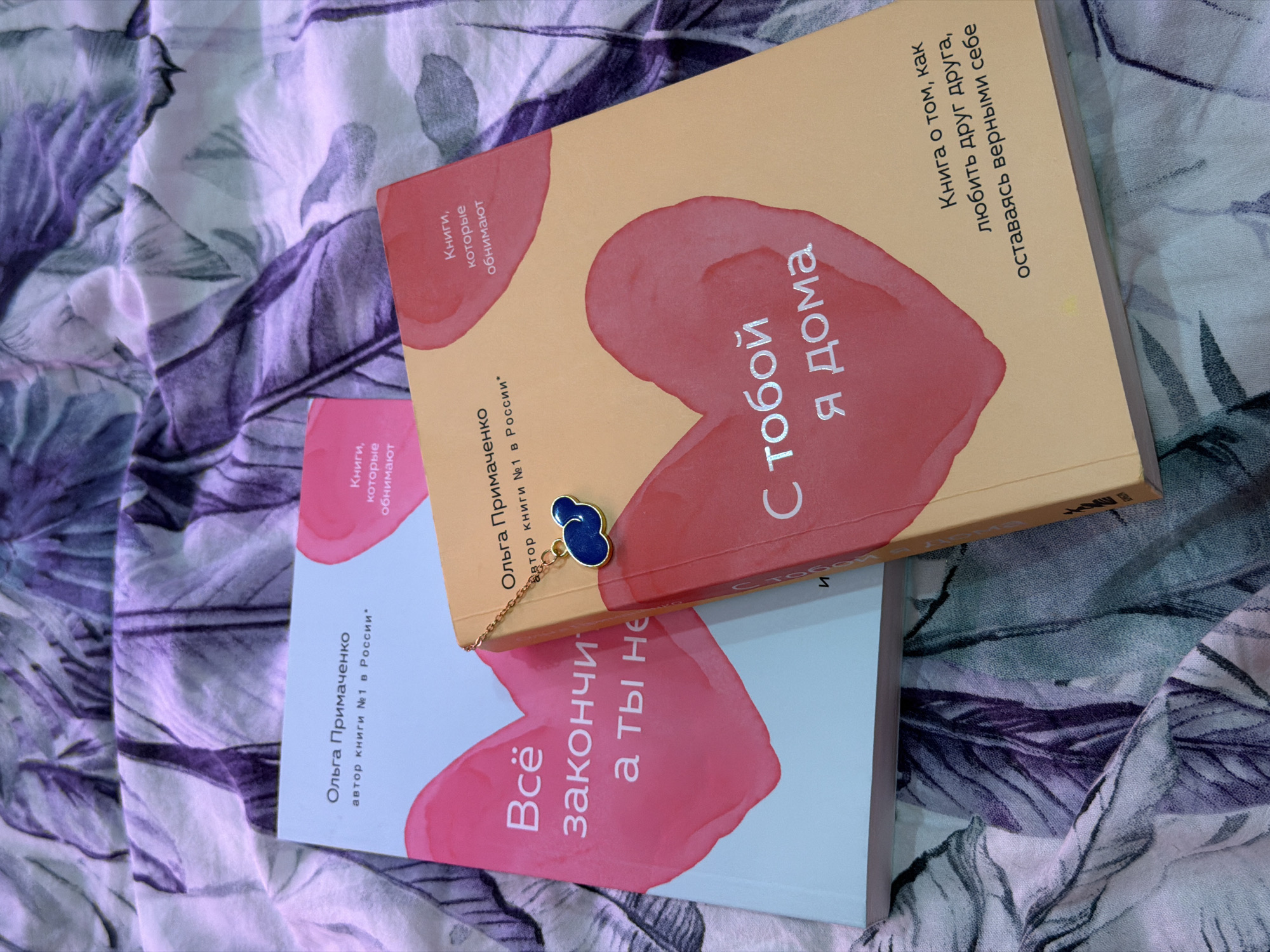 Here is a description of the image:
The image shows two books by Olga Primachenko stacked on a patterned surface. The top book has a yellow cover with a red heart shape on it, and the title "С тобой я дома" (With you I am home). The bottom book has a light blue cover with a pink heart shape, and the title "Всё закончится, а ты нет" (Everything will end, but not you). Both books are labeled as "Книги, которые обнимают" (Books that hug) and state that Olga Primachenko is the "автор книги №1 в России" (author of book number 1 in Russia). A blue and gold charm is placed on the top book. The patterned surface has a floral or leafy design with purple and white colors. The location is not mentioned in the text and cannot be determined from the image.