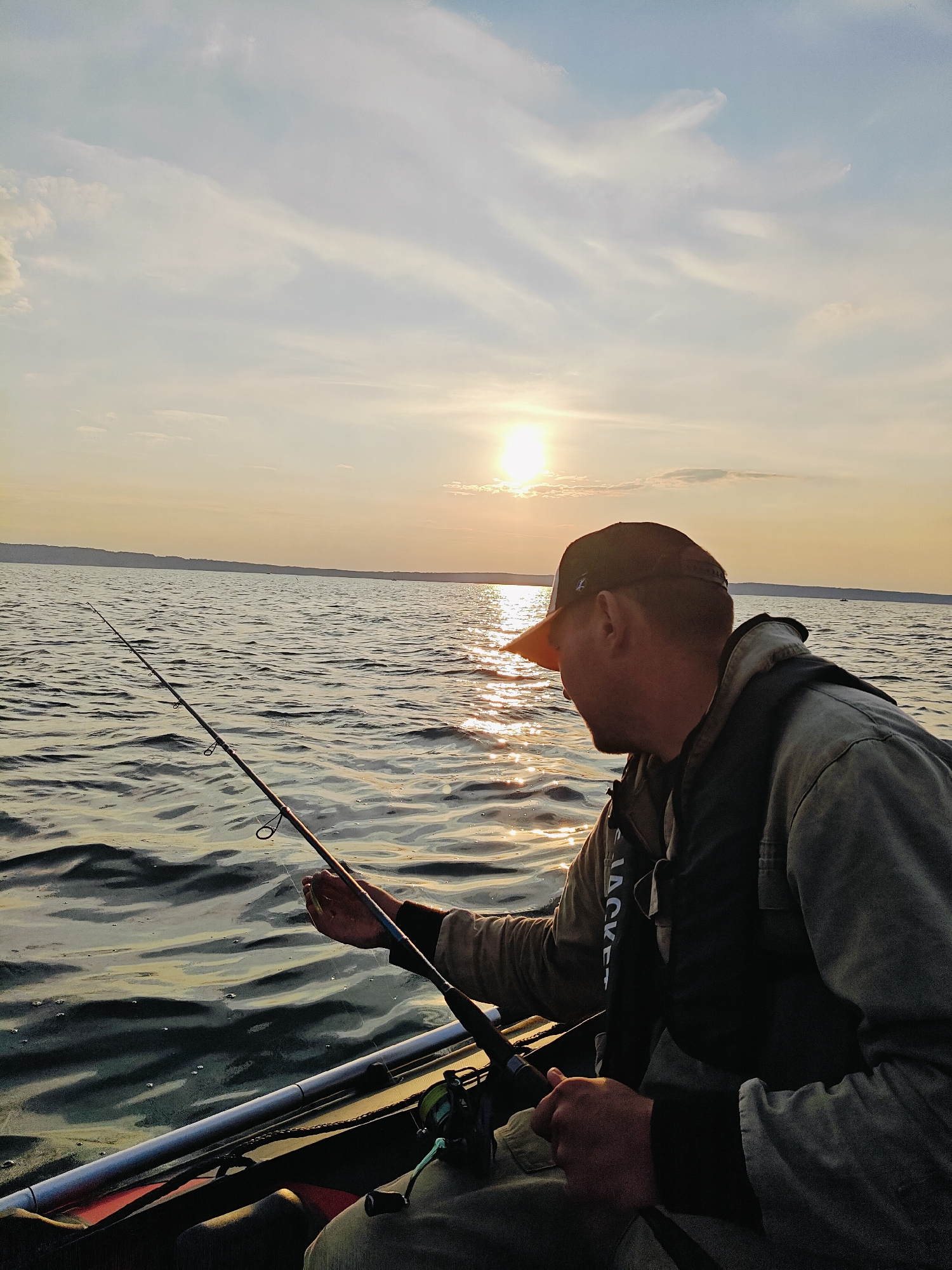 An adult male wearing a baseball cap and a life vest is fishing from a boat on a body of water. The sun is setting, casting a warm glow on the water and the sky. The man is holding a fishing rod and appears to be focused on his line. The water is rippling, and there is a shoreline visible in the distance. The scene is serene and suggests a peaceful evening spent outdoors. The location is the Kama River in Chaykovsky, Russia, known for its scenic beauty and recreational opportunities, including fishing.