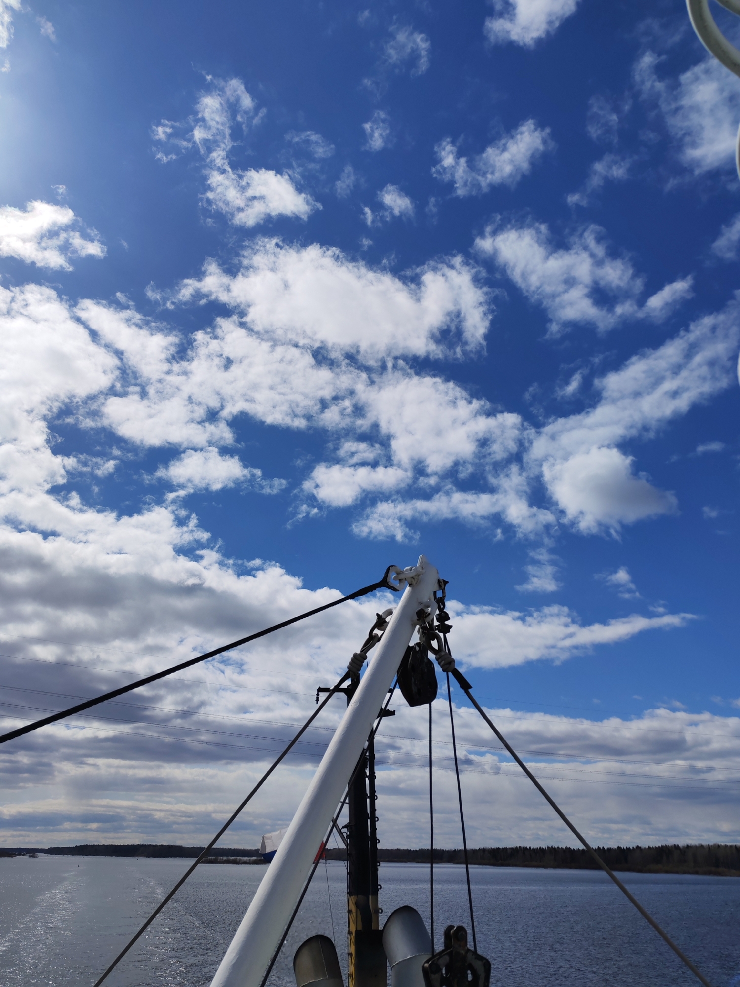The image shows a view from a boat or ship. A white mast with rigging is in the foreground. In the middle ground is water and land with trees. The sky is blue with scattered white clouds.