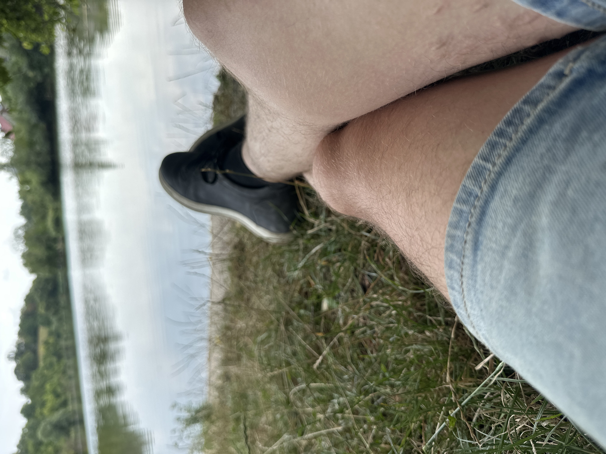 The image shows a close-up, low-angle shot of a person's legs and feet resting on the grassy bank of a body of water. The person is wearing denim shorts and dark-colored athletic shoes. The background consists of a calm lake with reflections of trees and the sky on its surface. The environment appears to be a natural setting, possibly a park or recreational area near Makiv, Ukraine. The focus is on the relaxed posture of the individual enjoying the scenery.