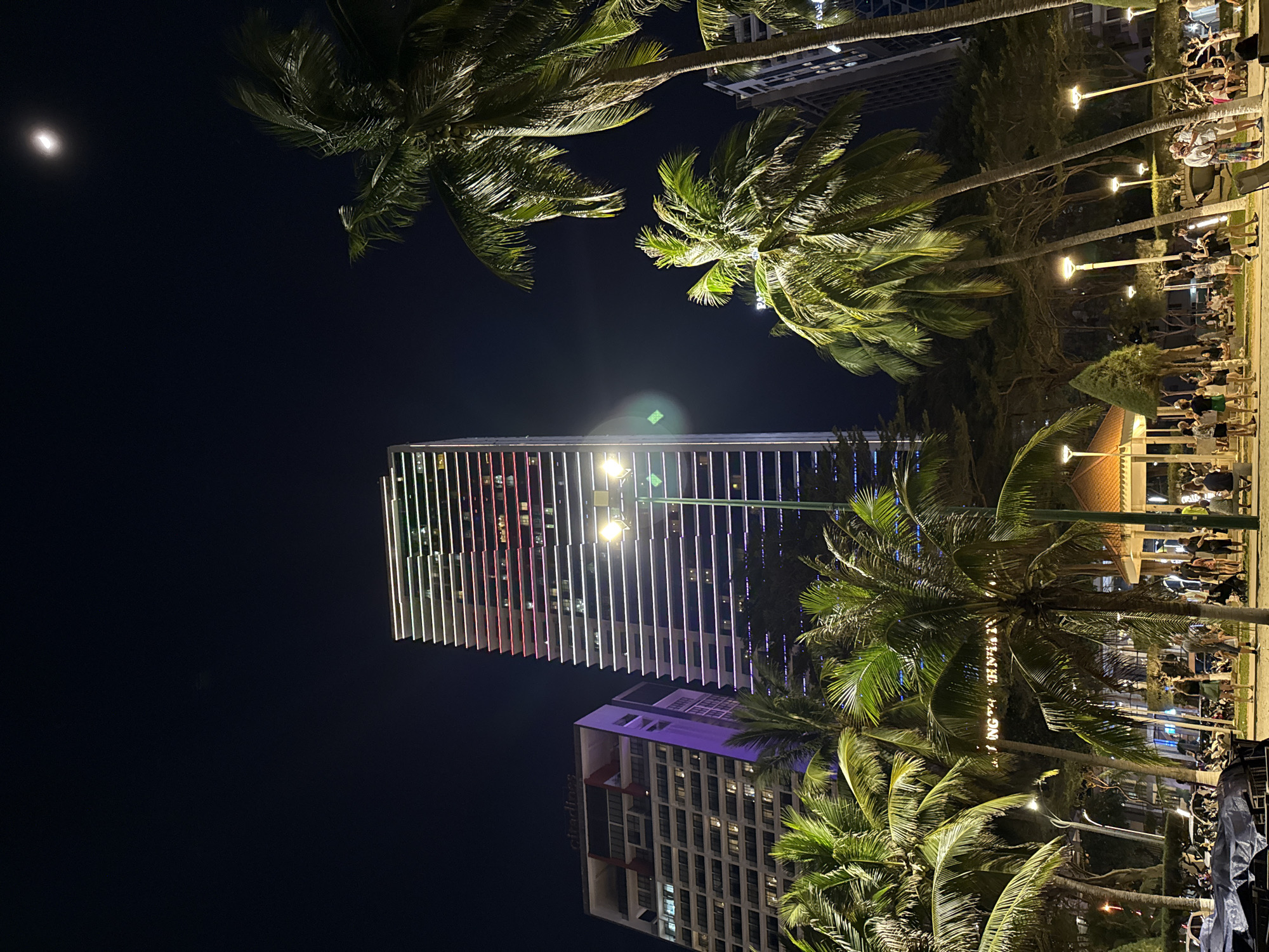 This image captures a vibrant night scene in Nha Trang, Vietnam, illuminated by a mix of natural and artificial light. The time of day is clearly night, with a dark, clear sky where a bright crescent moon shines in the upper left.
The central focus is a tall, modern skyscraper whose facade is strikingly lit with vertical stripes of white, red, and purple lights, creating a dynamic visual effect. Below it, another multi-story building is partially visible, featuring the discernible text "Alacarte" on its side, suggesting a hotel or commercial establishment.
Numerous lush palm trees frame the scene, their fronds glowing green from ambient artificial light sources. Some of these trees have visible coconuts. A strong light source, possibly a streetlight, creates a prominent lens flare near the illuminated skyscraper.
On the right side of the image, a lively gathering of people fills what appears to be a wide promenade or plaza. They are seen standing and strolling under the warm glow of numerous streetlights that line the area, suggesting an evening social event or leisurely activity. While individual faces are not distinct, their collective presence indicates an active atmosphere. In the bottom right corner, some parked vehicles, possibly motorbikes, are partially obscured by a tarp. Text is also visible wrapped around one of the palm trees, though largely illegible, it appears to contain Vietnamese words like "TÁNH NHỮNG".
The overall impression is one of a bustling tropical city evening, characterized by modern architecture, abundant greenery, and public enjoyment.