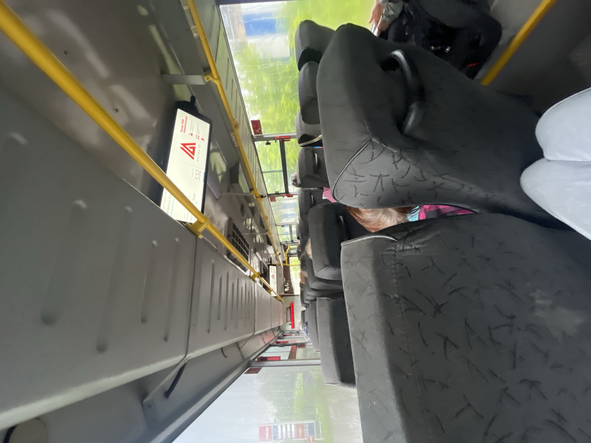 The image is taken from the interior of a bus, looking forward along the aisle. The seats are upholstered in dark gray fabric with a subtle pattern. Yellow handrails run along the ceiling. A digital display above the seats shows route information, including the number "532" and text that appears to be in Russian. Outside the windows, a blurry, green, outdoor environment is visible, suggesting a route passing through a somewhat vegetated area, possibly on the outskirts of a city. In the distance, a red and white sign is visible through the left window, possibly a roadside advertisement. The lighting is overcast, suggesting it may be daytime but not particularly sunny. The image appears to be taken in Krasnokamsk, Russia, based on the prompt.