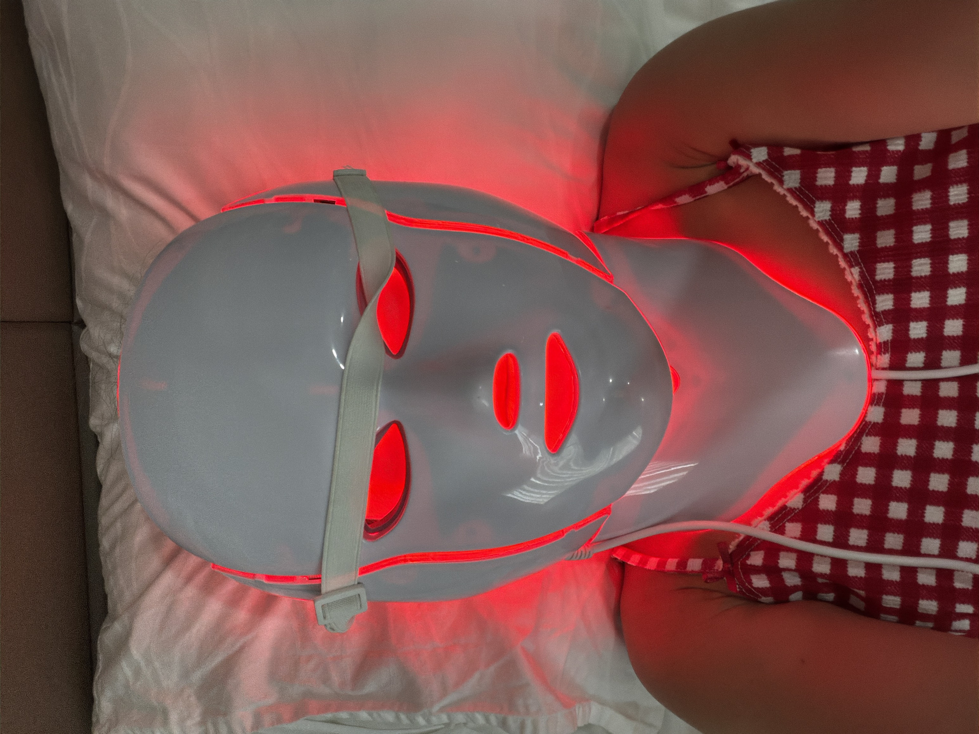 A person is lying down wearing a white LED facial mask that emits red light. The mask covers their entire face, with openings for the eyes and mouth. The red light emanates from within the mask, creating a glow around the face and neck. The person is wearing a red and white checkered top. They are lying on a white surface, possibly a bed or a treatment table, which is partially covered by a white sheet. The lighting is predominantly red due to the mask, with some ambient light present. The image appears to be taken from a top-down perspective. The location in the city of Nha Trang, Vietnam is not determinable from the provided image.