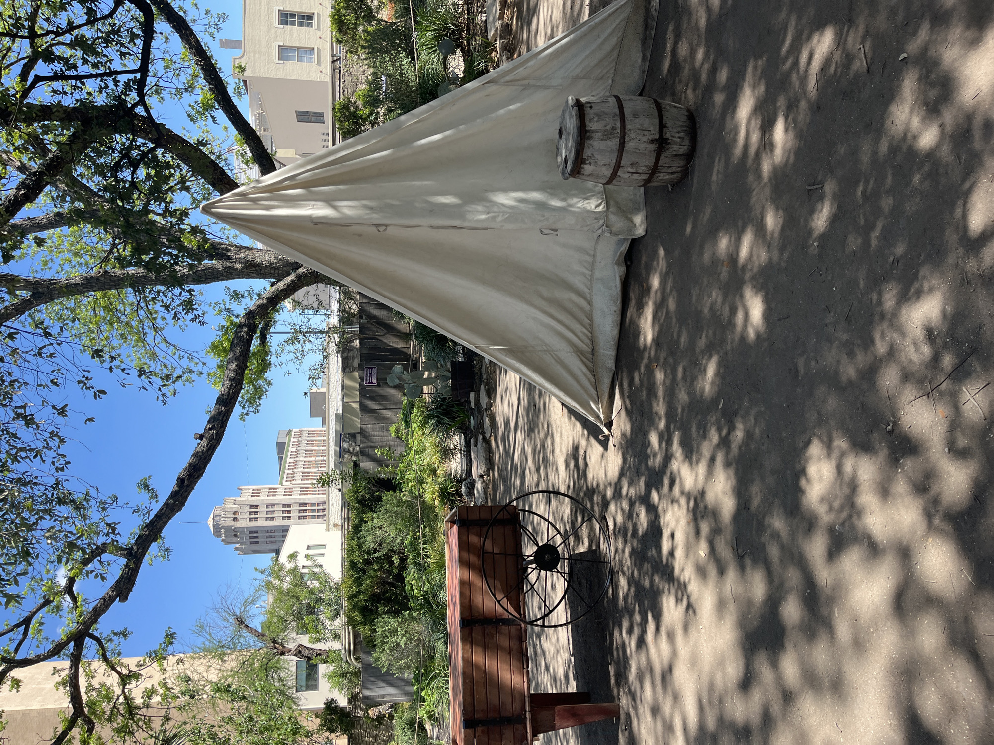 The image shows an outdoor scene in San Antonio, Texas. A white canvas tent stands on the right, with a wooden barrel placed next to it. A wooden cart with a large metal wheel is positioned nearby. The ground is dirt and is covered with shadows. In the background, buildings are visible including the Tower Life Building and other architecture. A tree branch with green leaves is visible in the upper left of the image. The sky is blue and clear.