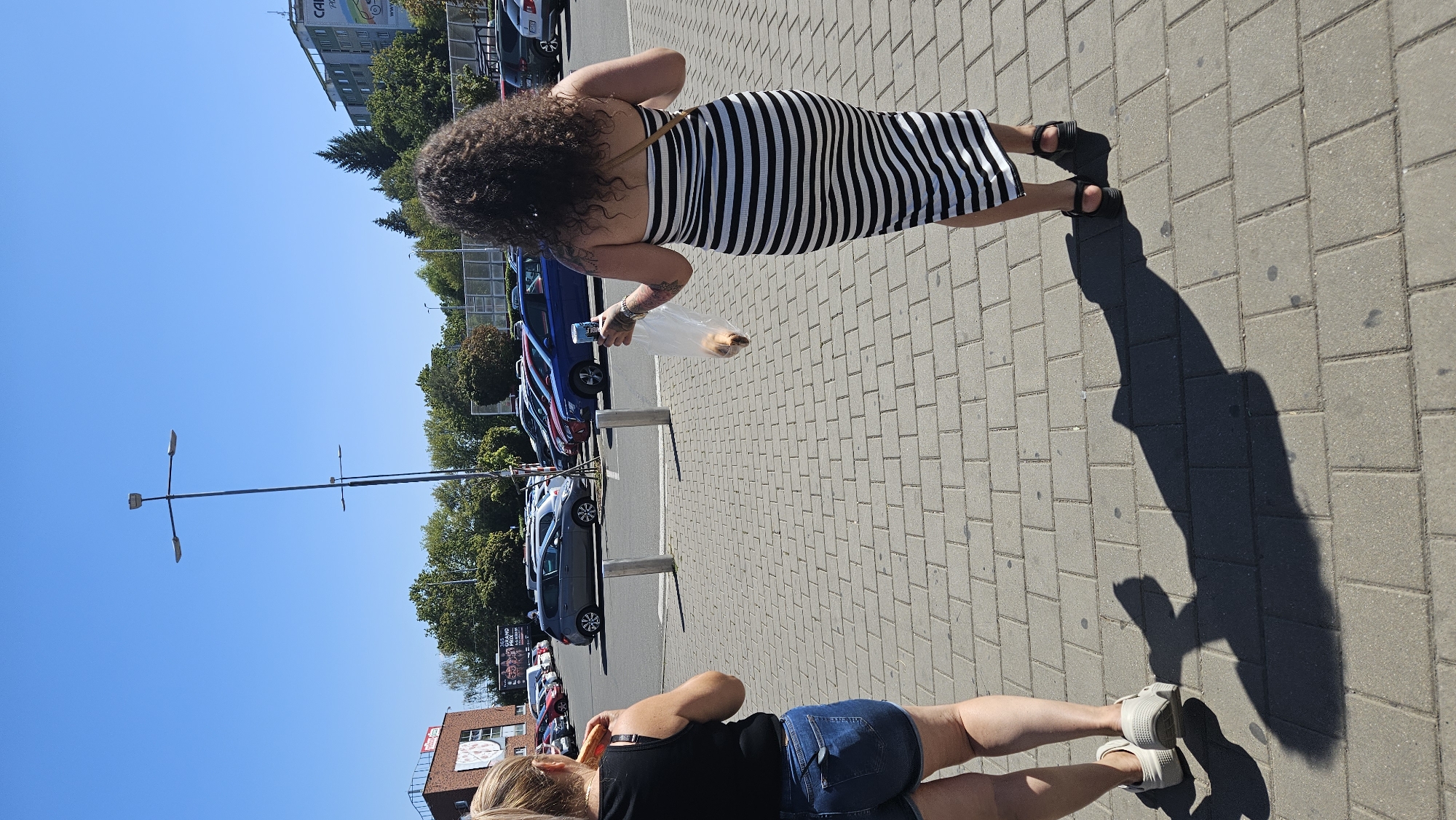 The image shows two women walking on a paved sidewalk in a city. The woman on the left, facing away from the camera, is wearing a black tank top and denim shorts and is eating a sandwich. The woman on the right, also facing away, is wearing a black and white striped dress and carrying a clear plastic bag with what appears to be bread rolls inside. She is also holding a blue bottle. In the background, there is a parking lot with several cars and a building with a sign that reads "EUROVEA". There are also streetlights and trees visible. The sky is clear and blue. The scene appears to be in an urban environment, possibly a shopping center or a commercial area in Prešov, Slovakia, given the presence of a prominent building with an international name like "EUROVEA", although the specific location in Prešov cannot be confirmed solely from the image content.