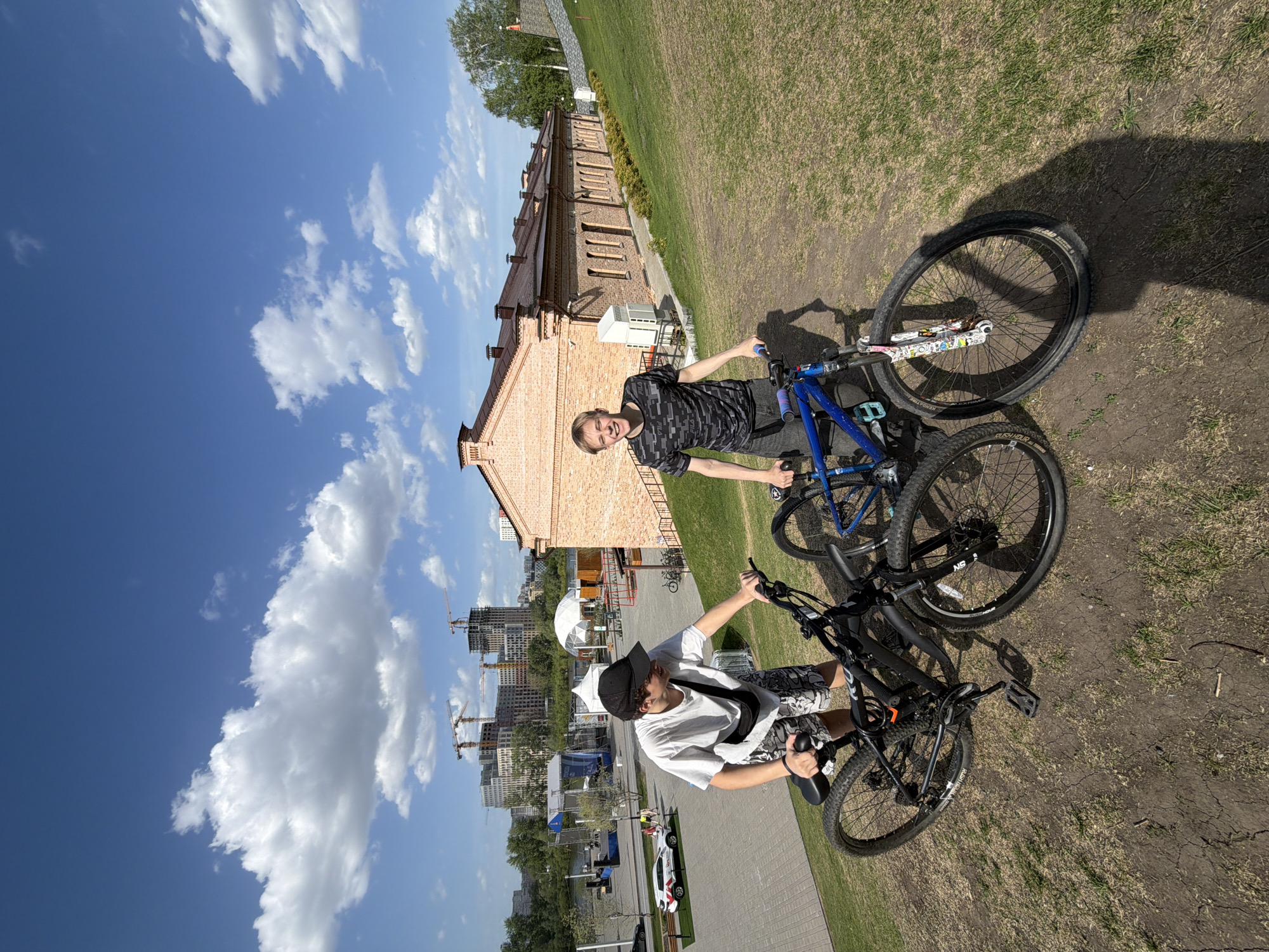 Two people with bikes are standing in front of the "History Park Russia - My History" in Tyumen, Russia. One person is wearing a black and grey shirt and riding a blue bike. The other person is wearing a white shirt, shorts with a black pattern and a black baseball cap. Behind them is a red brick building. In the background there are tall buildings and construction cranes. The sky is blue with white clouds. They appear to be on a patch of dirt near some grass and a paved sidewalk.