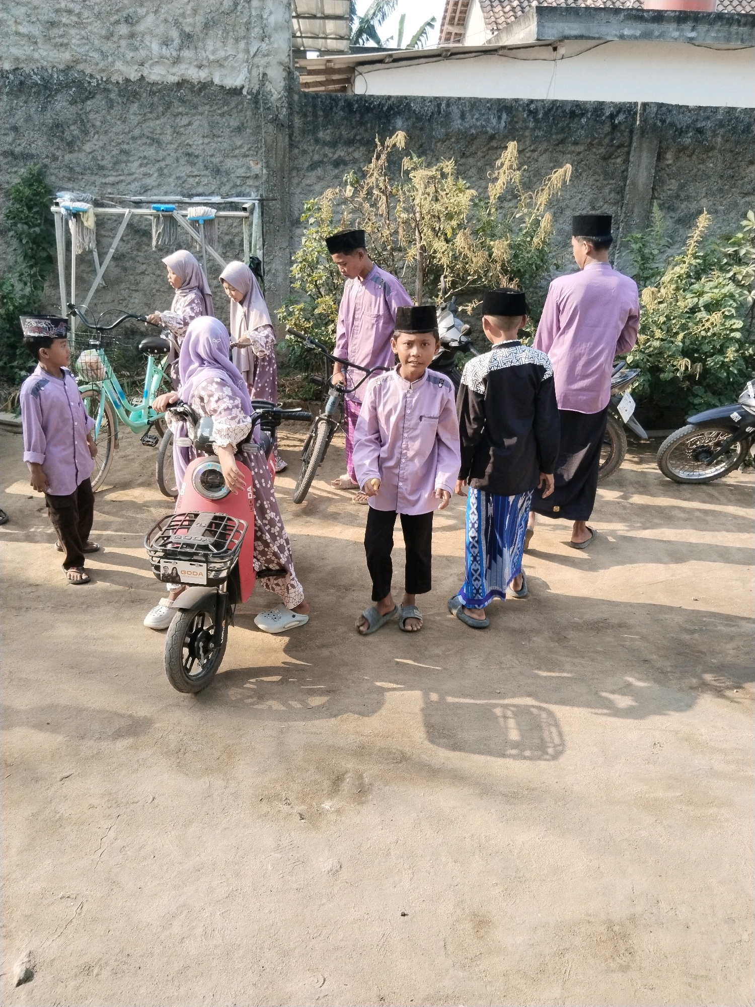 The image depicts a group of people, including men and children, in an outdoor environment. The scene appears to be in a residential area, with a concrete wall in the background and a dirt ground. Several individuals are dressed in traditional Muslim attire, specifically "baju koko" (a type of shirt) and "sarong" (a wrap-around skirt), and are wearing "peci" (a cap). There are also bicycles and a small red electric scooter present in the image. One child is seated on the electric scooter, while others stand around it. The lighting suggests it is daytime, likely morning or afternoon, with shadows cast on the ground. The location is identified as Gedung Meneng, Indonesia.