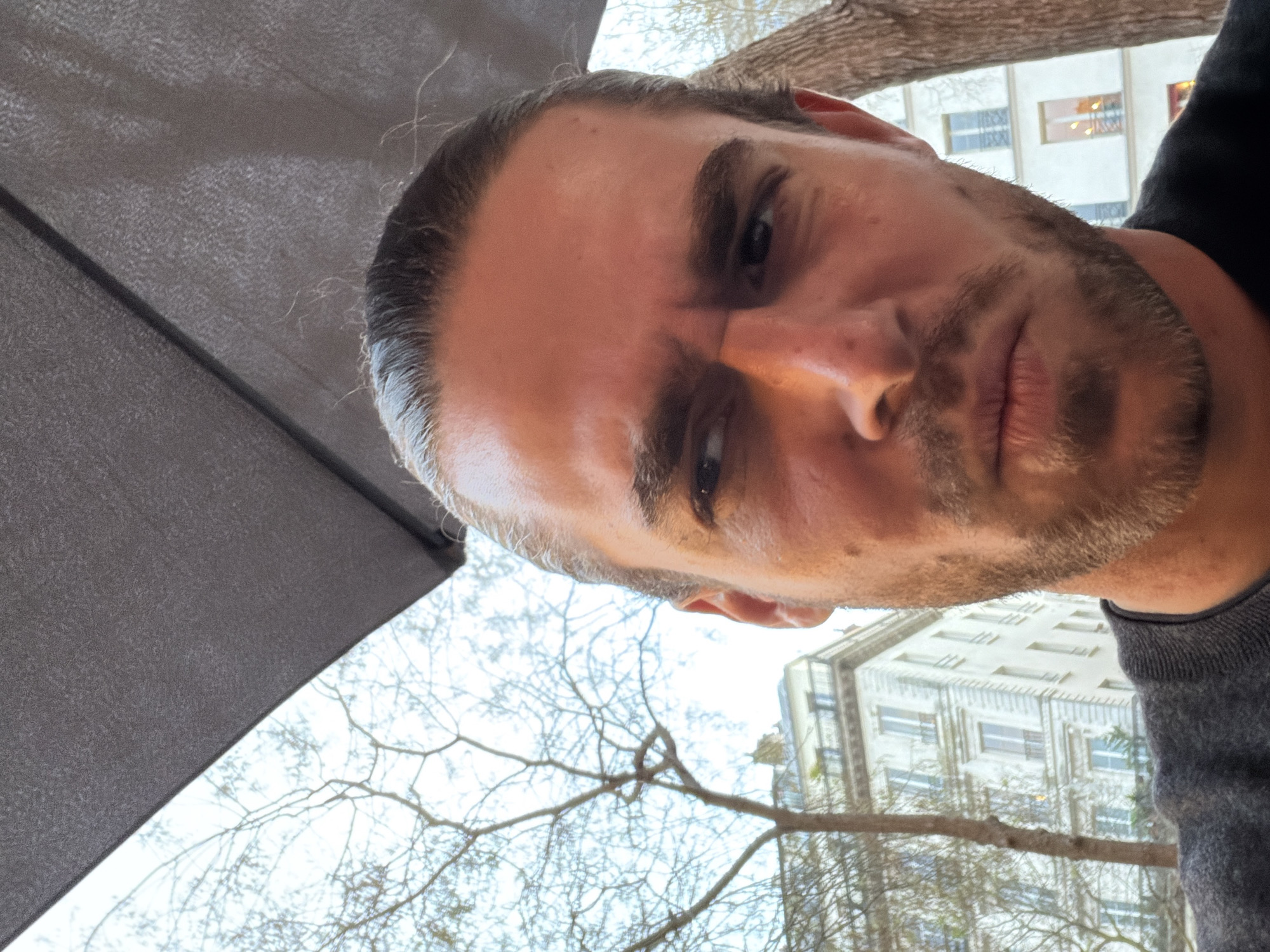 A close-up portrait of a light-skinned male with short brown hair pulled back is captured from a low angle. The subject has a light brown beard and is wearing a grey sweater and a black shirt. He is looking slightly downward with a slight frown. In the background, a building with multiple windows can be seen, along with bare tree branches. A grey, umbrella-like structure is visible on the left side of the frame. The presence of the building and architectural style suggests a possible location in Paris, France.