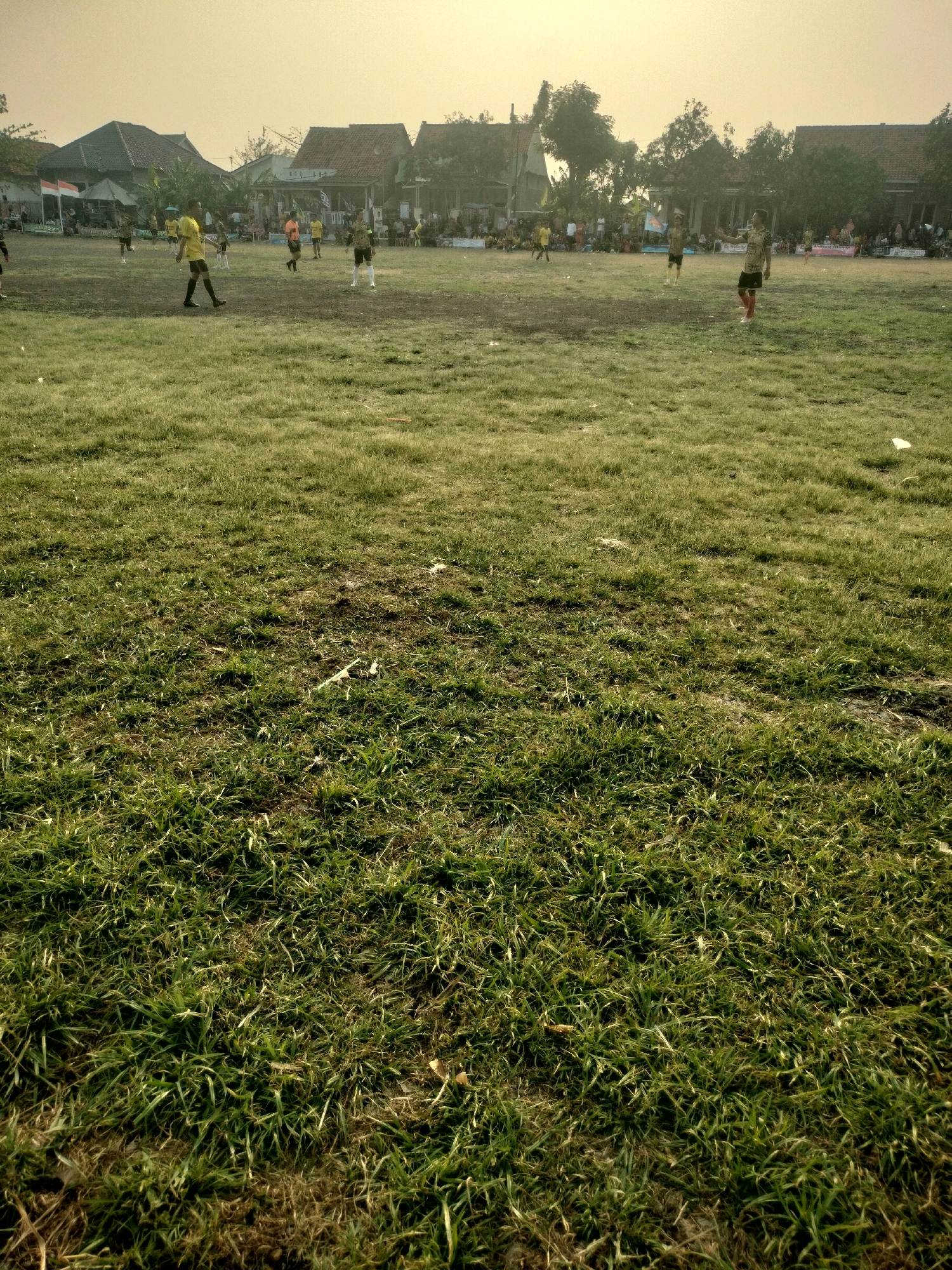 The image depicts an outdoor scene on a grass field, likely a soccer field, under a hazy sky. Several individuals are visible, some appearing to be players in sports attire, standing and walking on the field. In the background, there are buildings with red tile roofs, suggesting a residential or village setting. The presence of spectators lining the field indicates an ongoing event, possibly a match or practice. The ground is covered with grass, with some patches of bare earth and scattered debris. The overall atmosphere appears to be that of a community gathering or sporting event in a rural or semi-urban Indonesian environment. The location is identified as Kalisari, Indonesia.
