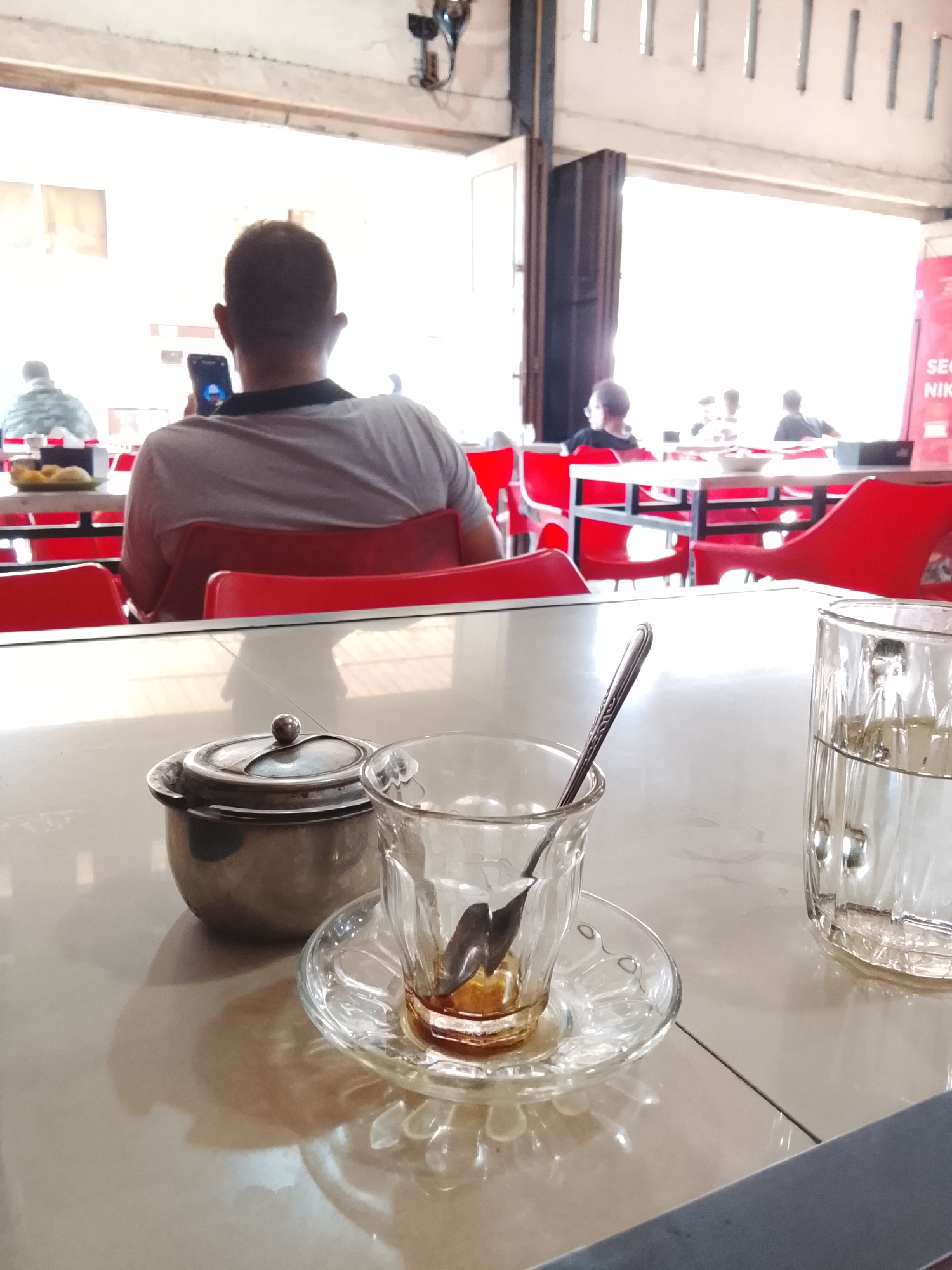 In a brightly lit open-air cafe in Lhokseumawe, Indonesia, the foreground features a table with a silver sugar bowl, a glass cup with remnants of coffee and a spoon inside, and a glass of water. A man sits facing away from the camera, looking at his phone. Further back, other patrons sit at red tables and chairs. The cafe is open to the outside. A red sign is visible on the right.