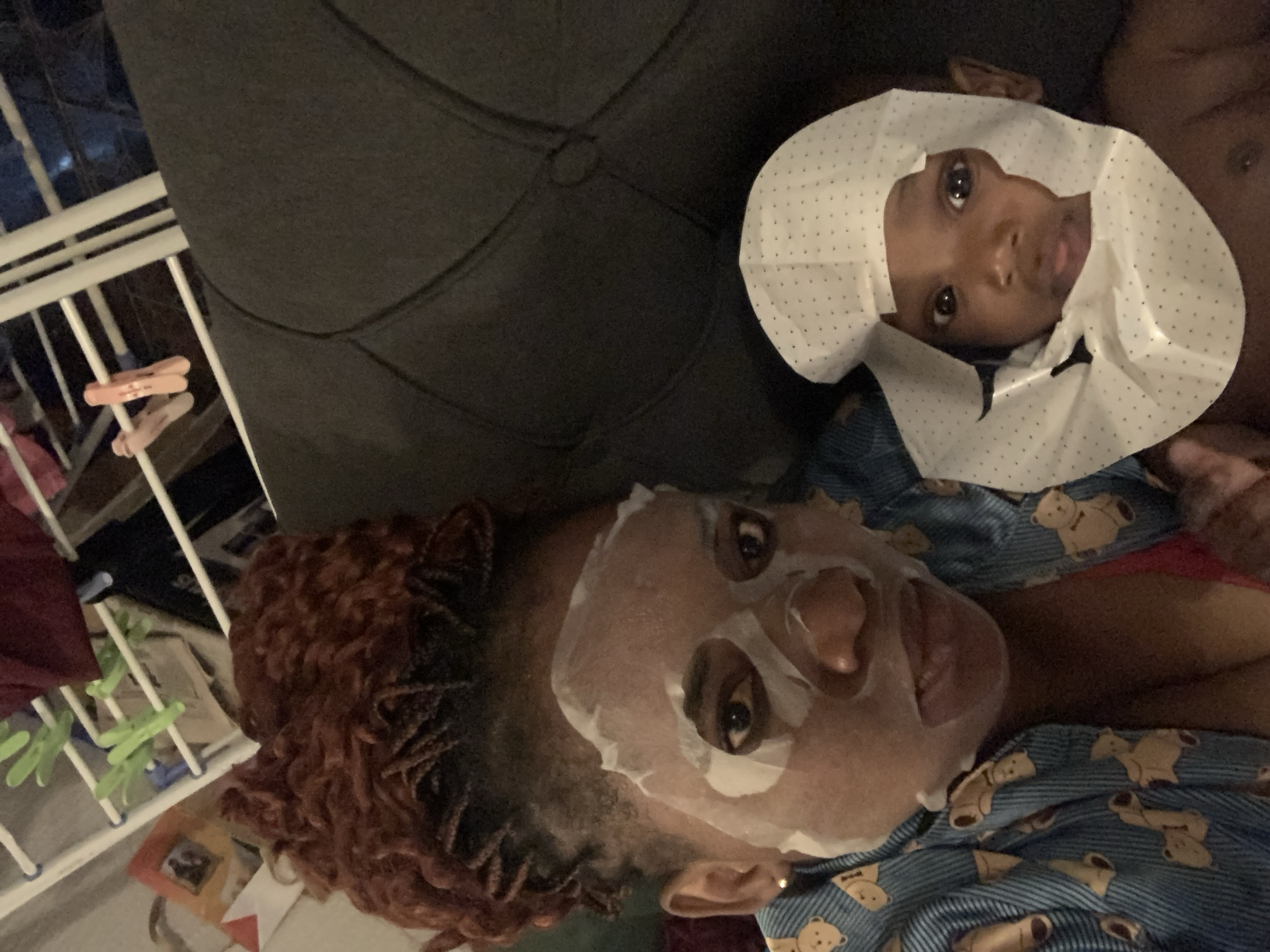The image presents a woman wearing a facial mask and lying next to a baby who is wearing a dotted white shower cap. The woman's hair is reddish-brown, styled in small braids or twists. She is wearing a blue pajama shirt with a teddy bear print. The setting appears to be indoors, with a glimpse of a clothes rack in the background. The location is not discernible as Barakin Akawo, Nigeria from the image alone.