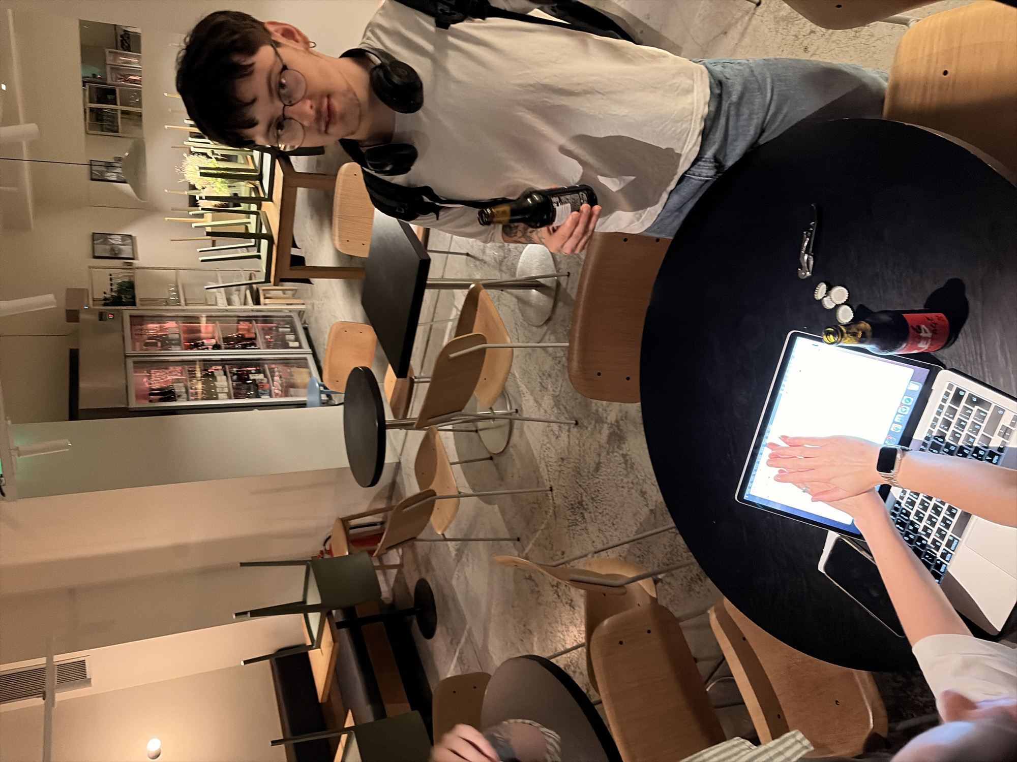 A man with glasses and headphones around his neck holds a dark glass bottle of beer, standing in what appears to be a bar or restaurant. He is wearing a white t-shirt and blue jeans. In the foreground, a laptop is open on a black round table, with a person's hands on the keyboard and a phone nearby. There are several other tables and chairs scattered around the room, some of which are stacked. A refrigerated display case filled with bottles is visible in the background, along with shelves of glassware. The environment suggests a casual setting, possibly a bar or cafe after closing hours, given the stacked chairs. The location is identified as Belgrade, Serbia.