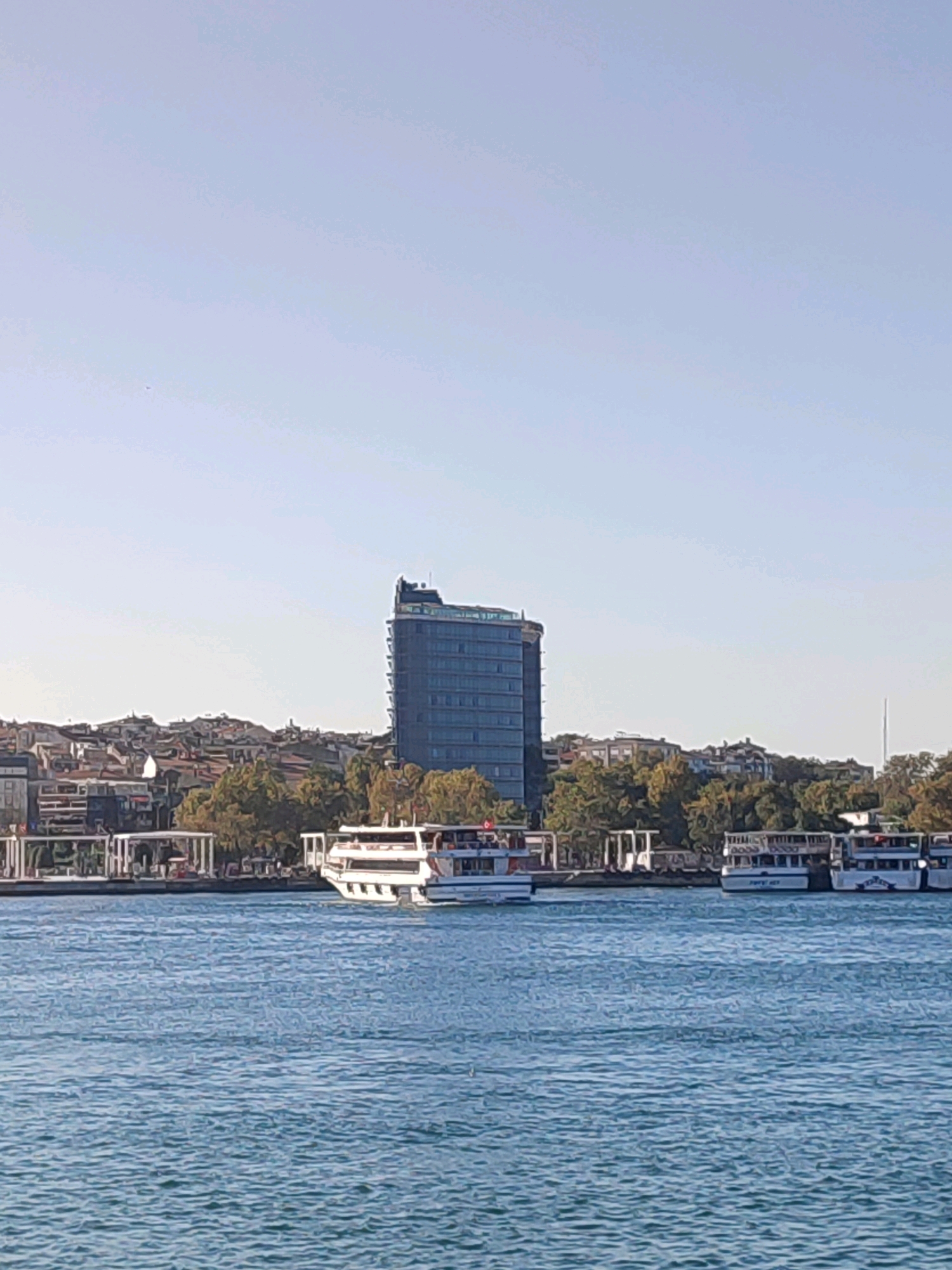 The image captures a wide view of a city skyline bordering a body of water, likely the Bosphorus Strait, near Kadıköy, Turkey. In the foreground, the blue water is textured with small ripples. Along the shoreline, several white ferries are docked or moving. A prominent modern glass-fronted skyscraper stands tall behind a dense cluster of traditional buildings with red-tiled roofs, characteristic of urban Turkish architecture. Lush green trees dot the landscape between the buildings and the water. The sky is a clear, pale blue with no visible clouds, suggesting a bright, sunny day. The overall scene conveys a bustling urban environment integrated with maritime activity.