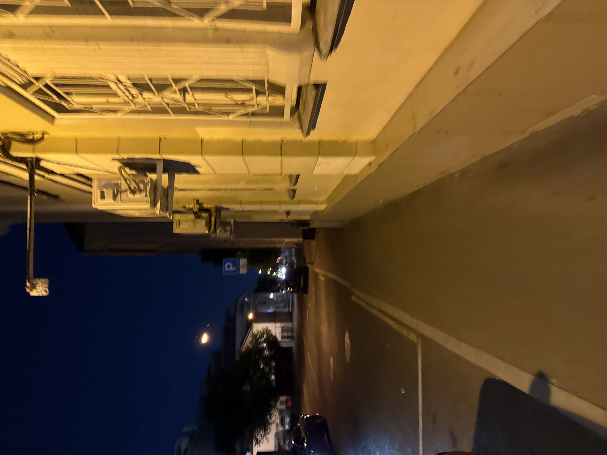 The image displays a nighttime urban street scene captured from a low-angle pedestrian perspective, looking forward along a sidewalk. The sky is a dark, deep indigo blue, indicating late evening or night.
On the immediate right, a multi-story building dominates the frame, its facade uniformly bathed in a warm, yellowish-orange light originating from unseen streetlights or building illumination. The building exhibits architectural details including vertical white pilasters or divisions that segment the yellow wall. Each visible window is fitted with an intricate, multi-paned decorative security grate, presenting a complex geometric pattern. Several white, rectangular external air conditioning units are mounted prominently on the facade between windows, with visible conduits connecting them. Below the windows, dark blue or grey sills are present. Near the top right of the building, a small, white, rectangular traffic signal unit is attached to a horizontal arm.
The foreground consists of a dark grey sidewalk, also illuminated by warm light, leading into the distance. A distinct white line delineates the sidewalk's edge from the dark asphalt street, and a thinner yellow line runs along the curb. A soft shadow is cast on the lower left portion of the sidewalk.
The street, paved with dark asphalt, curves gently to the left in the mid-distance. A dark-colored vehicle is partially visible on the far left of the street, its headlights casting a faint glow. Further down the street, another dark vehicle is discernible, along with distant vehicle lights. A single, bright streetlight on the left side of the street illuminates a section of the road ahead. A standard blue square "P" parking sign, featuring a white capital letter "P," is mounted on a pole on the right side of the street, positioned further from the viewer. Across the street, dark silhouettes of trees with dense foliage are interspersed with glimpses of less illuminated buildings. The background consists of the dark, featureless night sky, devoid of stars or other distinct celestial objects, with distant ambient city lights subtly marking the urban expanse.
This scene is characteristic of an older, central district street in Moscow, Russia, featuring typical urban architecture, street infrastructure, and artificial lighting found within the city's established areas.