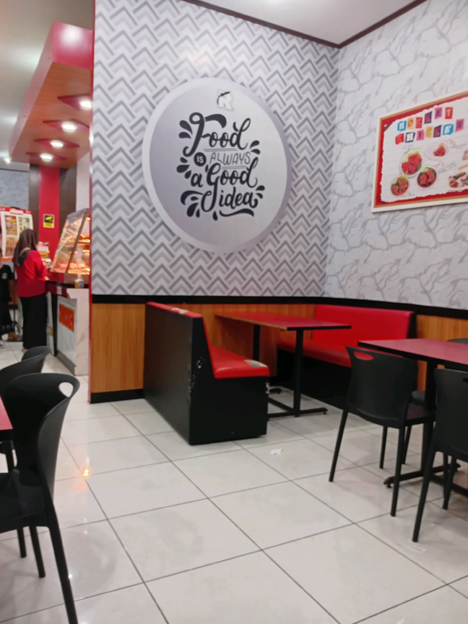 The image shows the interior of a fast-food restaurant in Pejagoan, Indonesia. The dining area features red booth seating and black chairs around red-topped tables. The walls are decorated with a geometric pattern and a large circular graphic with the text "Food Is Always A Good Idea." A framed menu or advertisement is visible on the right wall. In the background, a glass display case, likely for food items, is present, and a woman in a red top and black pants is standing near it, facing away from the camera. The floor is tiled in white with gray grout lines. The overall ambiance suggests a clean and modern fast-food establishment.
