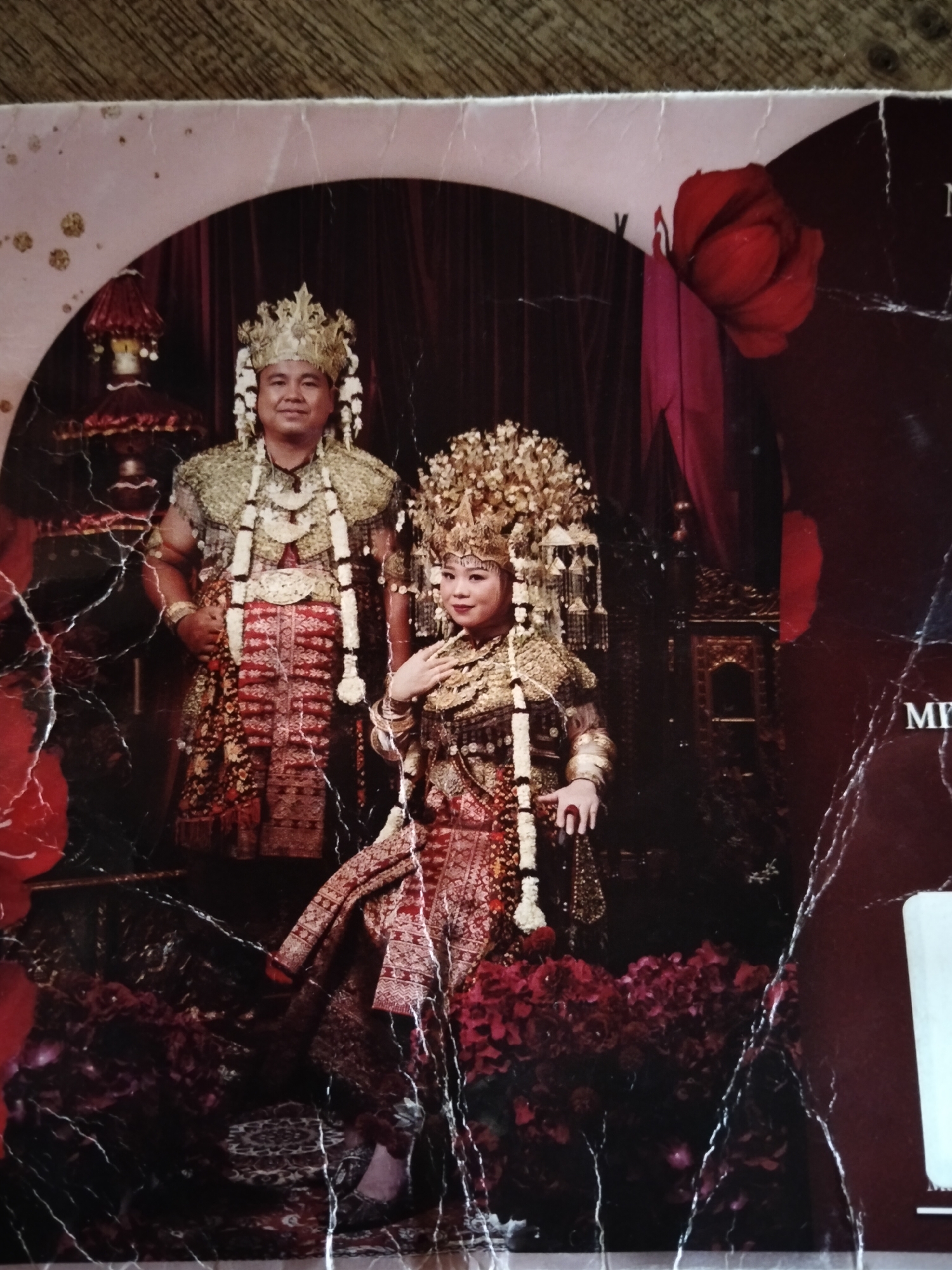 This is a photograph of a couple in traditional Indonesian wedding attire, possibly from the Minangkabau culture of West Sumatra. The man on the left wears a golden headdress, a patterned vest, and a red, patterned sarong. He has a necklace with white beaded tassels. The woman on the right wears an elaborate, tall golden headdress adorned with decorative elements, a matching golden ornate top, and a red, patterned sarong. She also wears a necklace with white beaded tassels and her hands are adorned with rings and bracelets. They are posed in front of a dark, ornate background with red drapery and what appears to be intricate furniture or architectural elements. There are floral arrangements in deep red hues in the foreground. The photograph itself shows signs of damage, with cracks and creases across the surface.