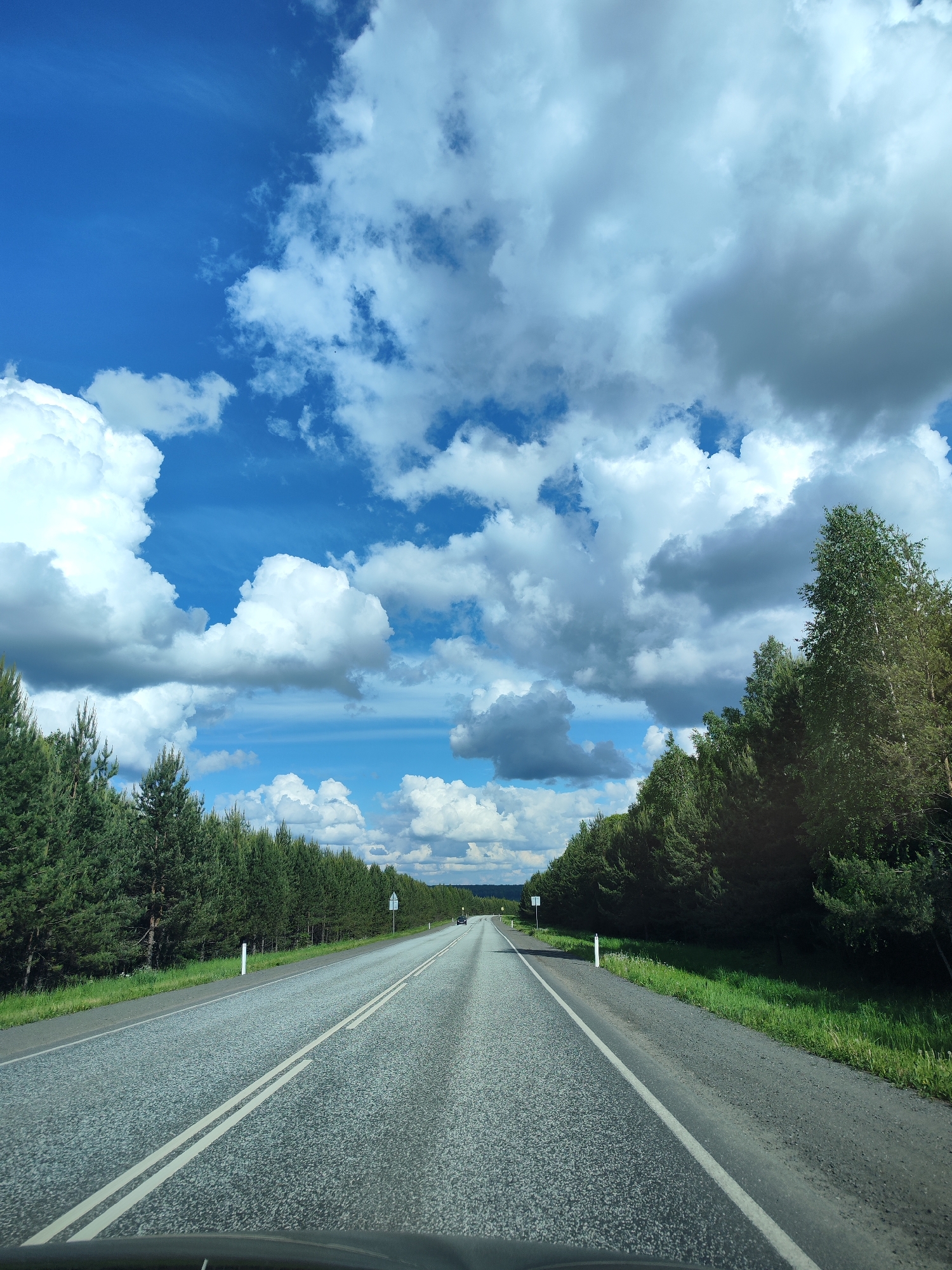 The image depicts a straight, asphalt road bisecting a landscape in Сосново, Russia. The road is marked with double white lines down the center and is bordered on both sides by dense green forests. The sky above is predominantly blue, scattered with large, puffy white clouds, some of which have grey undersides. The scene suggests a rural or semi-rural setting, with the road appearing to be the main thoroughfare. There is no visible human activity.