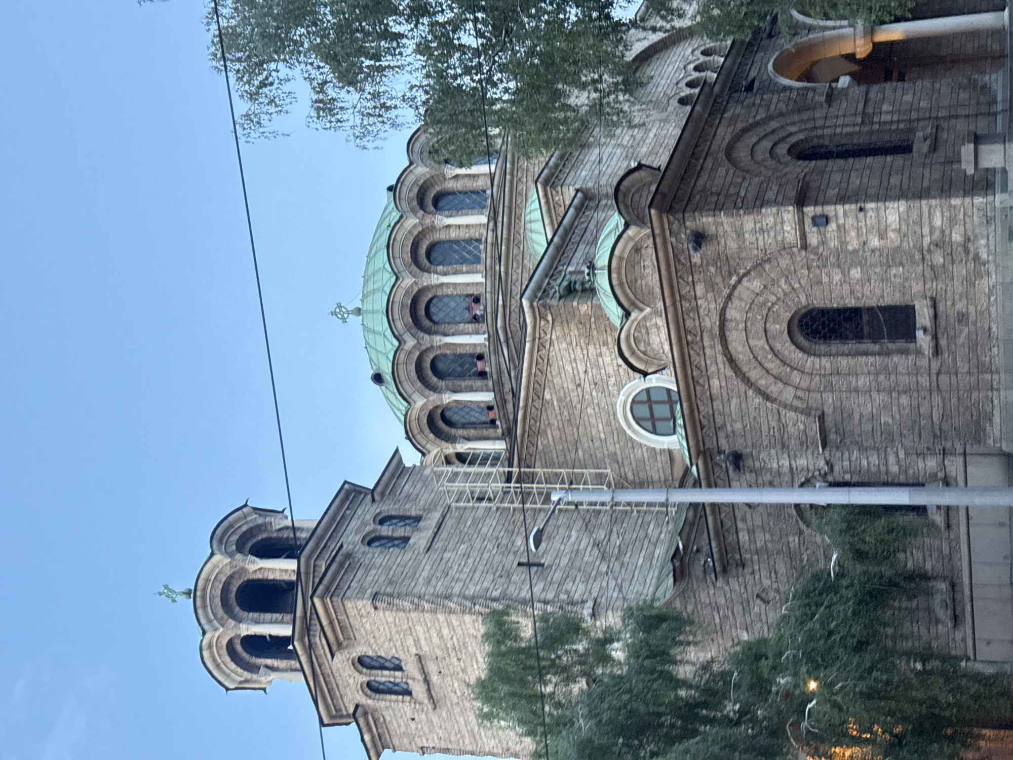 This image captures a low-angle view of the Alexander Nevsky Cathedral in Sofia, Bulgaria. The imposing stone structure is bathed in the soft light of what appears to be either dawn or dusk, with a clear blue sky overhead.
The cathedral features a prominent bell tower on the left, topped with a copper-green dome and a cross. To the right, the main body of the cathedral is visible, dominated by a large, similarly colored dome, also adorned with a cross. Both domes are surrounded by smaller arched windows. The architecture showcases intricate stonework with arched windows and decorative elements across its facade.
In the foreground, the branches of several trees with lush green foliage partially obscure the lower sections of the building. A utility pole with a street lamp stands to the left of the bell tower. Electrical wires stretch across the scene, connecting to unseen points. In the background, the sky is a gradient of pale blue. While the image shows the exterior of the cathedral, there are no visible people in the frame. The scene is static and conveys a sense of architectural grandeur.