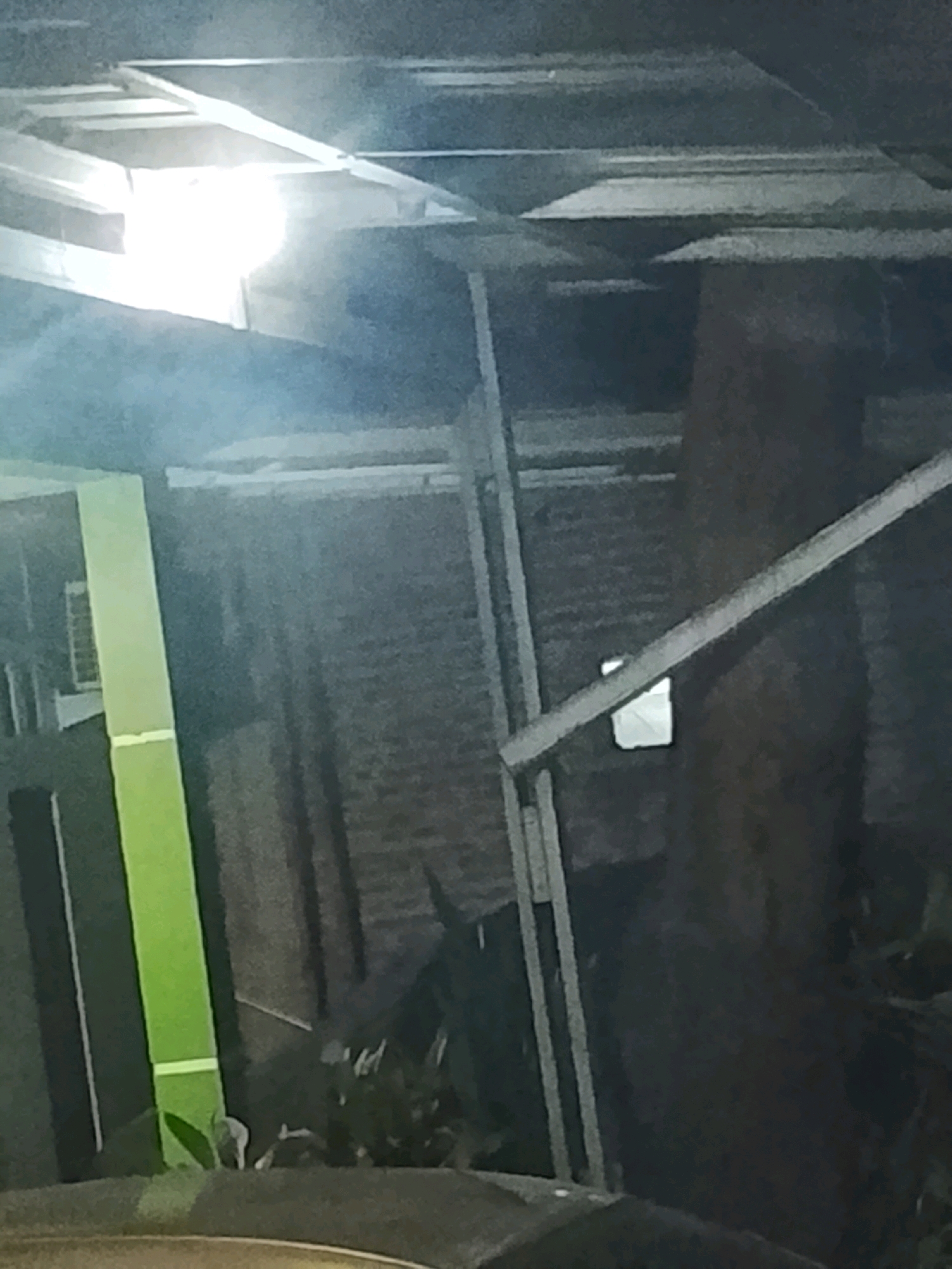 The image captures a dimly lit scene, possibly at night, featuring an outdoor structure with a corrugated metal roof. To the left, a tall, lime green pillar stands prominently against a dark background that appears to be a brick wall. The pillar has white horizontal lines near its base. In the upper left portion of the image, a bright light source illuminates a section of the metal roof, creating a glare. A diagonal wooden beam runs across the right side of the frame, with a small, bright rectangular object visible beneath it. The overall environment suggests an area with some construction or an unfinished building. The scene is devoid of any people. The location is identified as Ujungrusi, Indonesia.