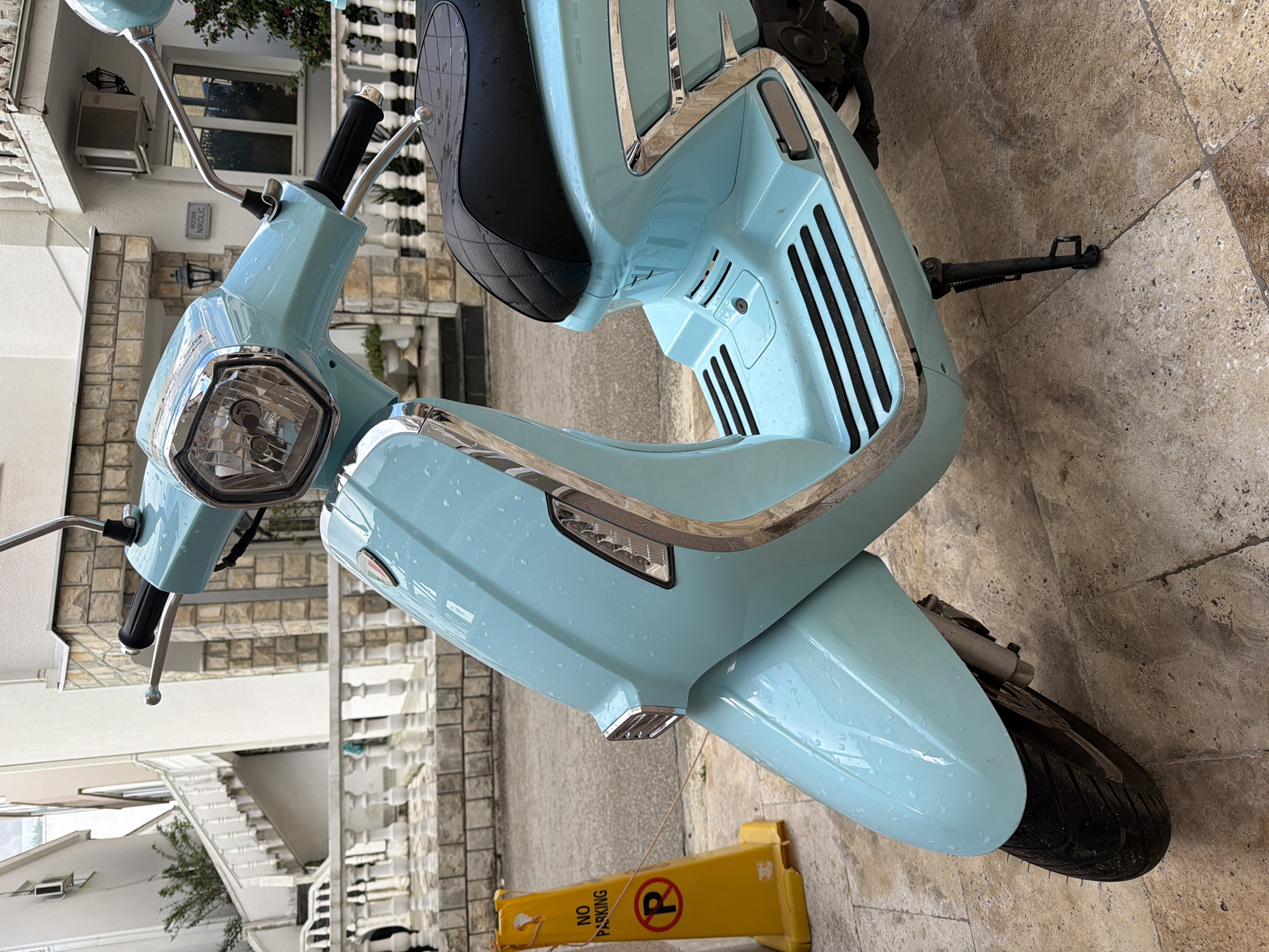 A close-up, medium-shot photograph shows a light blue Lambretta scooter with chrome detailing, resting on a tiled surface in Budva, Montenegro. The scooter is oriented towards the left of the frame, and a yellow "No Parking" sign is near its front wheel. The environment includes residential architecture in the background, featuring stone walls, windows, and a white balcony. The scooter appears to be parked on a pedestrian walkway or patio. The photo gives a clear view of the scooter's design and its immediate surroundings.