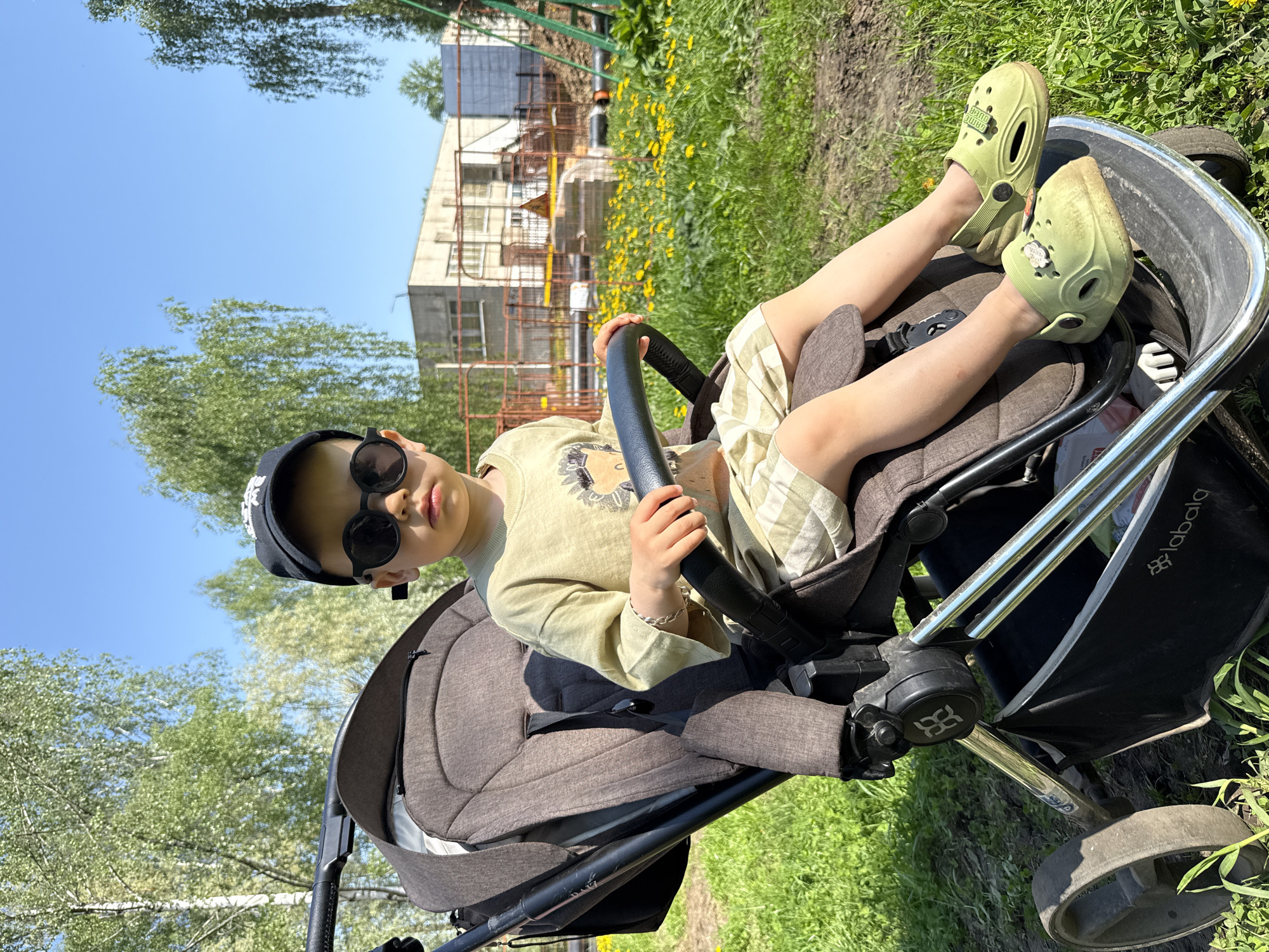 A young child, wearing a black baseball cap, sunglasses, a light green T-shirt, and striped shorts, sits in a stroller in an outdoor environment. The child holds a black circular object resembling a steering wheel. The child's feet are clad in light green Crocs. The stroller, which has a dark brown seat and a black frame, is positioned on grass next to a dirt path. In the background, there is a building with yellow flowers growing in front of it, and trees can be seen around the stroller. The image captures a moment in what appears to be a park or recreational area. The brand name "labala" is visible on the side of the stroller. This is Rusanovo, Russia.