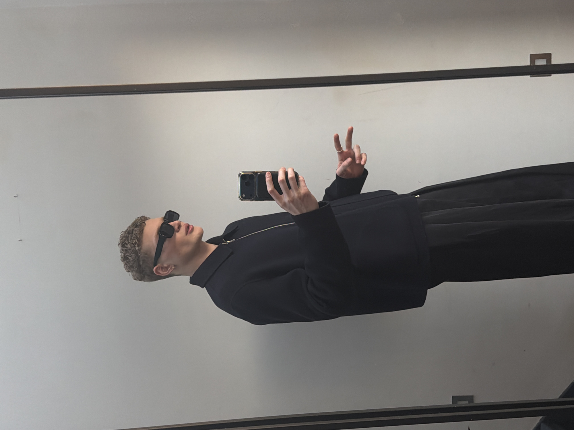 The image shows a man taking a selfie in a mirror. He has short, curly blonde hair and is wearing dark sunglasses, a black jacket, and black pants. He is holding a smartphone in one hand and making a peace sign with the other. The background is a plain gray wall with a dark frame around the mirror. Based on the style and ambiance, the setting could potentially be in London, United Kingdom, but there are no concrete landmarks or identifiers in the image to confirm this definitively.
