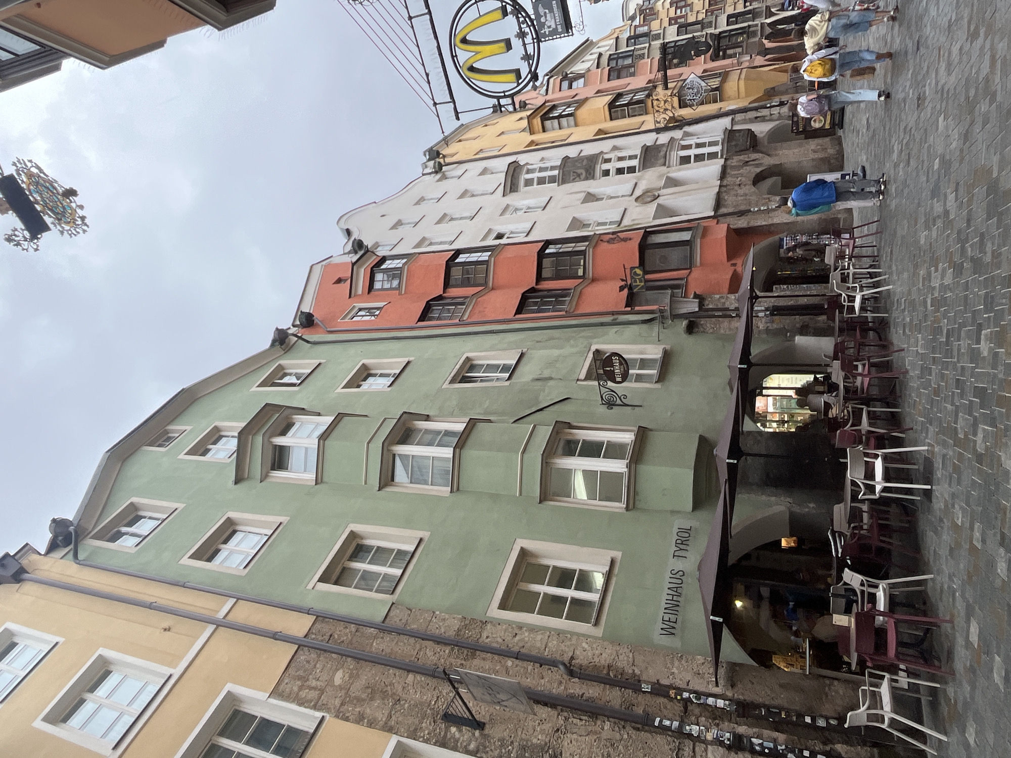 The image shows a street scene in Innsbruck, Austria, specifically the narrow alley of the historic Old Town. The buildings are tall and narrow, painted in vibrant colors such as yellow, green, coral, and white, with distinctive architectural features like bay windows. The street is paved with cobblestones. There are outdoor seating areas for restaurants and cafes with tables and chairs. A sign for "WEINHAUS TYROL" is visible on a green building. Further down the street, a McDonald's sign is prominently displayed. Several people are walking along the street, some carrying backpacks and shopping bags. The sky is overcast, suggesting a cloudy day.