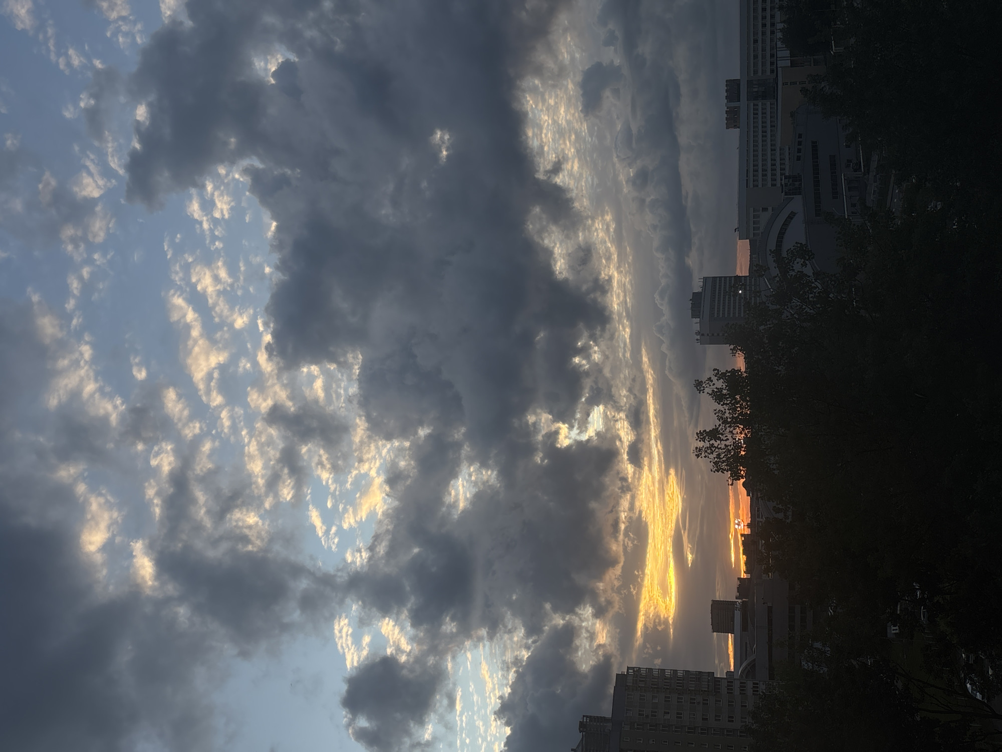 The image depicts a sunset over the city of Minsk, Belarus. In the foreground, the silhouettes of buildings are visible, partially obscured by trees. The sky is filled with clouds, ranging from dark gray to light blue, with the setting sun casting an orange and yellow glow through the gaps between the clouds. The perspective is from a ground-level vantage point, looking upwards towards the sky.