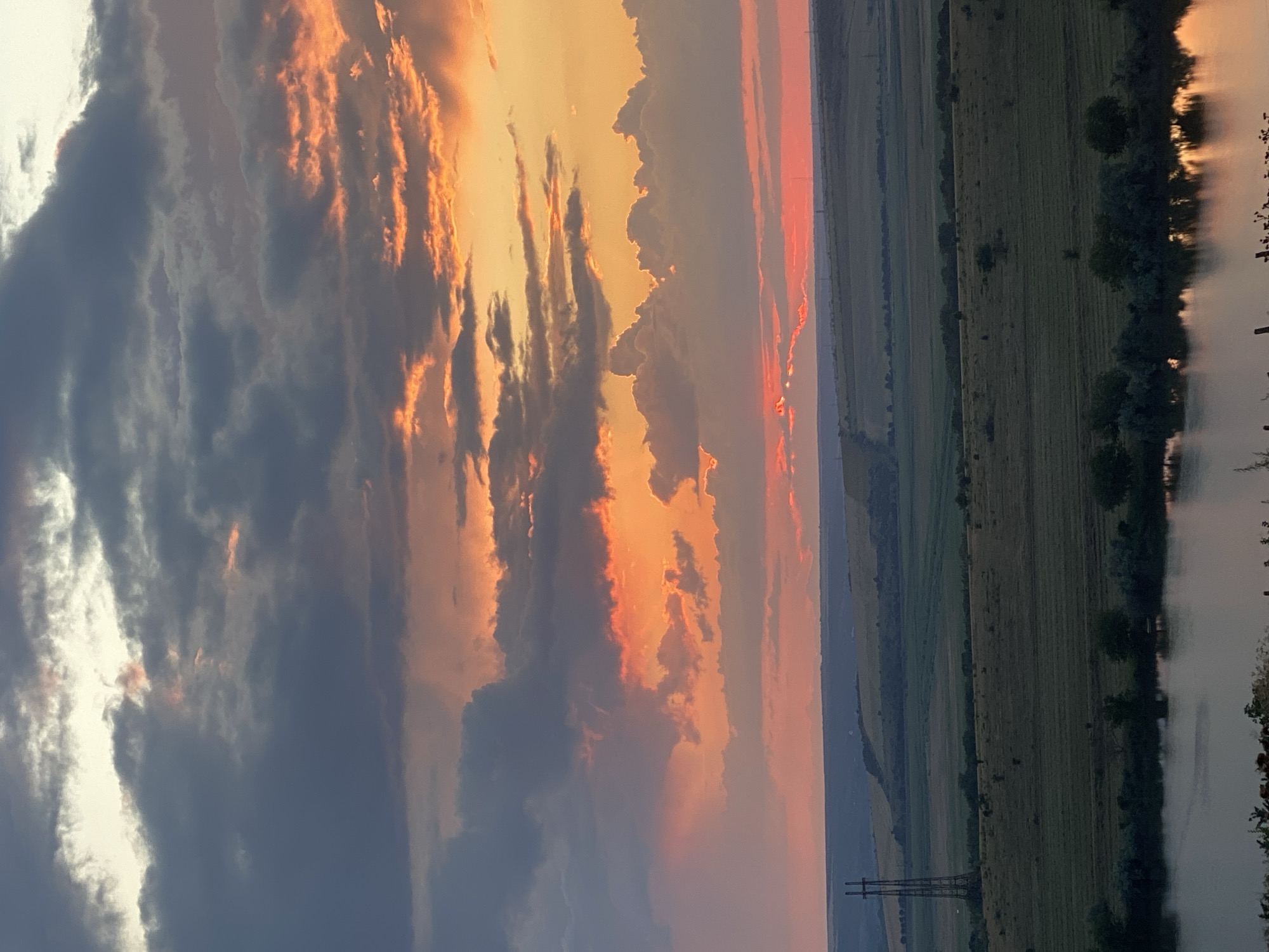 The image captures a dramatic sunset over a rural landscape, likely in Cairaclia, Moldova. The sky is filled with a gradient of colors, from deep grays and blues in the upper clouds to vibrant oranges, pinks, and yellows closer to the horizon. Wispy clouds are illuminated by the setting sun, creating a dynamic and textured sky.
Below the sky, rolling hills and fields stretch out, appearing as shades of green and brown. A body of water, possibly a lake or river, is visible in the foreground, reflecting the colors of the sky. Along the shoreline of the water, a line of trees and shrubs adds a dark, silhouetted contrast to the brighter sky. In the distance, a tall, latticed electricity pylon stands as a solitary man-made structure against the natural landscape, adding a touch of industrial presence to the otherwise serene scene. The overall atmosphere is one of natural beauty and tranquility, with the vivid sunset dominating the visual experience.