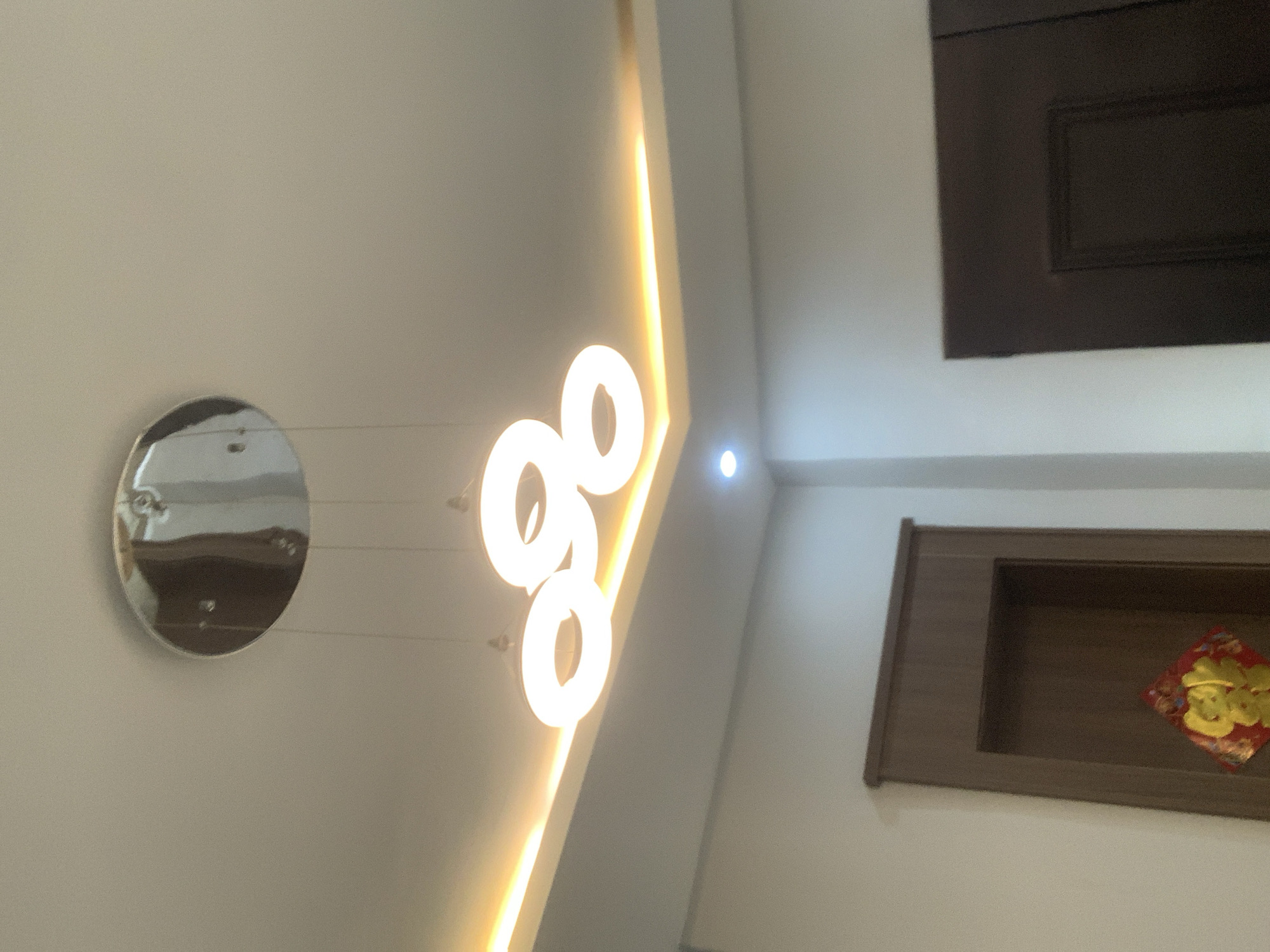 The image captures an interior view of a room, likely a home or a commercial space. The ceiling is the dominant element, featuring a modern recessed design with integrated LED strip lighting that emits a warm, golden glow. Suspended from the ceiling by thin wires is a contemporary chandelier composed of three interconnected circular light fixtures, also illuminated with a bright, white light.
To the left of the chandelier, a circular, reflective object, possibly a mirror or a decorative disc, hangs on the wall. Its surface shows distorted reflections of the surrounding environment, hinting at a dark, possibly wooden, surface.
On the right side of the image, a dark wooden niche is set into the wall. Within this niche, a small, red and gold ornament is visible, possibly a traditional decoration. The overall lighting suggests it is either daytime with the interior lights on, or evening. The scene appears calm and well-lit, with no people visible. The style of the decor suggests a modern aesthetic. The mention of Abuja, Nigeria, places the scene within this specific geographical context.