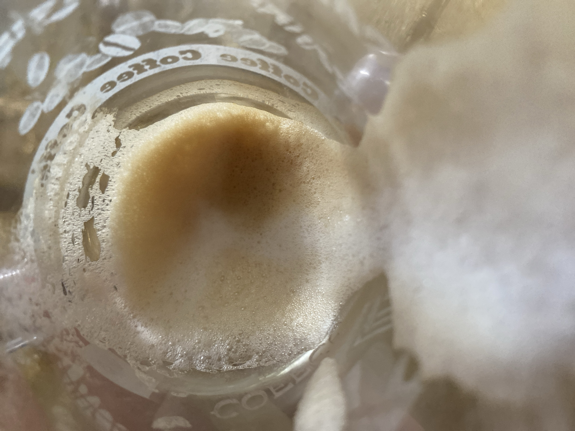 The image shows the inside of a glass with a foamy beverage, likely coffee, and the words "Coffee" printed around the rim. The beverage is a light brown color with a thick layer of foam on top. Some of the foam appears to be spilling over the side of the glass. The glass is transparent and shows reflections of light. There are also some coffee beans depicted in white around the rim of the glass. The image is taken from a top-down perspective, looking into the glass. There is no indication of the location being in the city of Maykop, Russia.