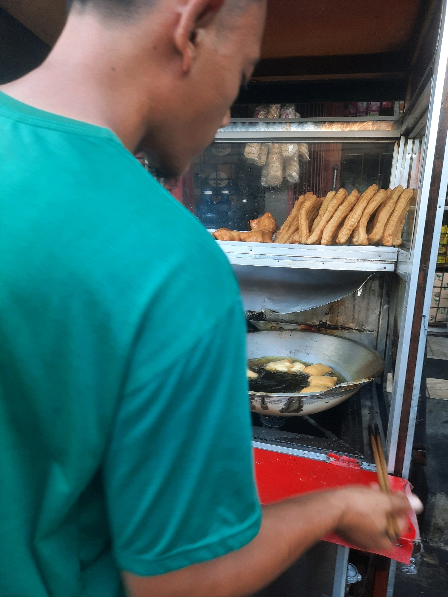A person wearing a turquoise t-shirt is standing next to a food cart in North Jakarta, Indonesia. The cart has a display case on top filled with fried dough sticks and some other fried items. Below the display case, there is a large wok filled with hot oil, where more pieces of fried dough are cooking. The person is holding a pair of chopsticks, appearing to be in the process of frying or handling the food. The background shows some shelves with packaged goods and bottles. The scene suggests a street food vendor preparing and selling fried dough.