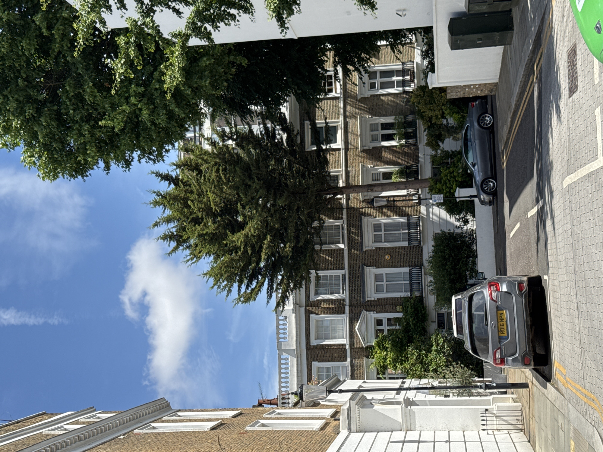 The image depicts a street scene in London, United Kingdom. The perspective is from above, looking down on a residential street. Dominating the foreground are the tops of trees with lush green foliage, obscuring parts of the view. Beyond the trees, a brick building with multiple windows and decorative white trim is visible. Several cars are parked along the street. One car, a dark gray sedan, is parked closer to the foreground, while a silver SUV with a yellow license plate is further down the street. The street is paved with gray cobblestones and has double yellow lines indicating parking restrictions. The sky is blue with a few scattered white clouds, suggesting a bright, sunny day. The architectural style of the buildings and the presence of parked cars on a residential street are characteristic of many London neighborhoods. There is no visible activity or people in the frame, creating a sense of stillness. The license plate on the silver SUV shows the text "RD16 NPN".