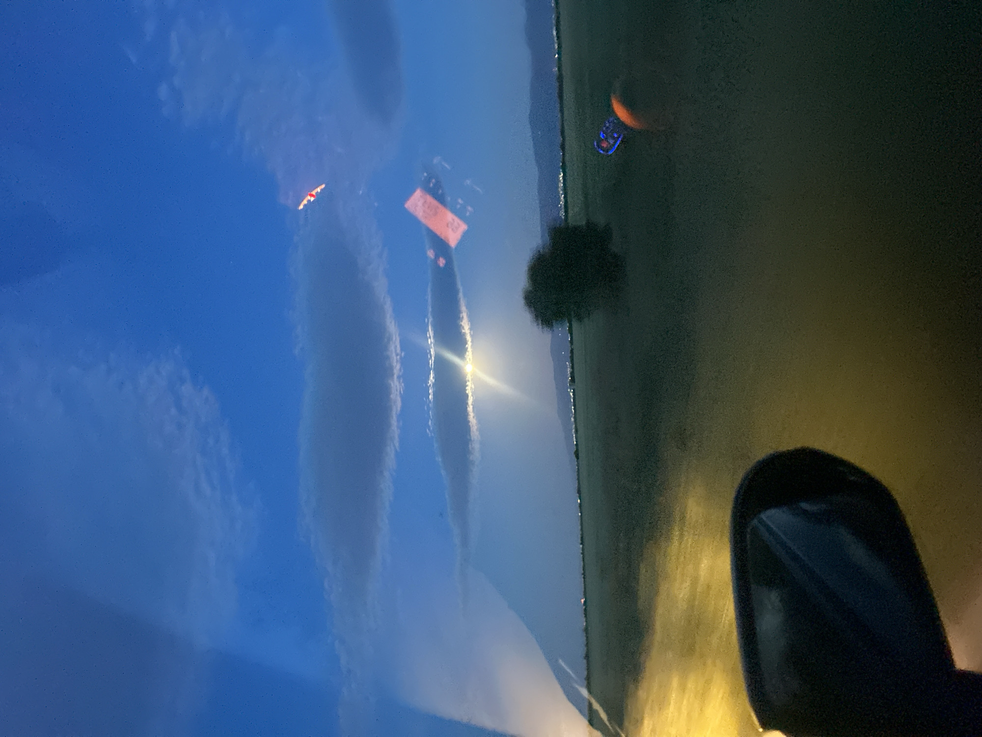 The image appears to be taken from inside a moving vehicle at dusk or night, with a view through the windshield. The left side of the image shows a reflection of the sky with light clouds, and possibly a bird or small aircraft. In the center of the windshield, there is a reflection of an orange sign. Outside the vehicle, there is a field or grassy area, with a tree visible near the center of the image. A vehicle side mirror is visible in the lower right corner, with the car's headlights illuminating the road. There are faint lights of what could be a city in the distance and a partial moon in the sky. The image does not provide specific clues about the location in Pishtigovo, Bulgaria.