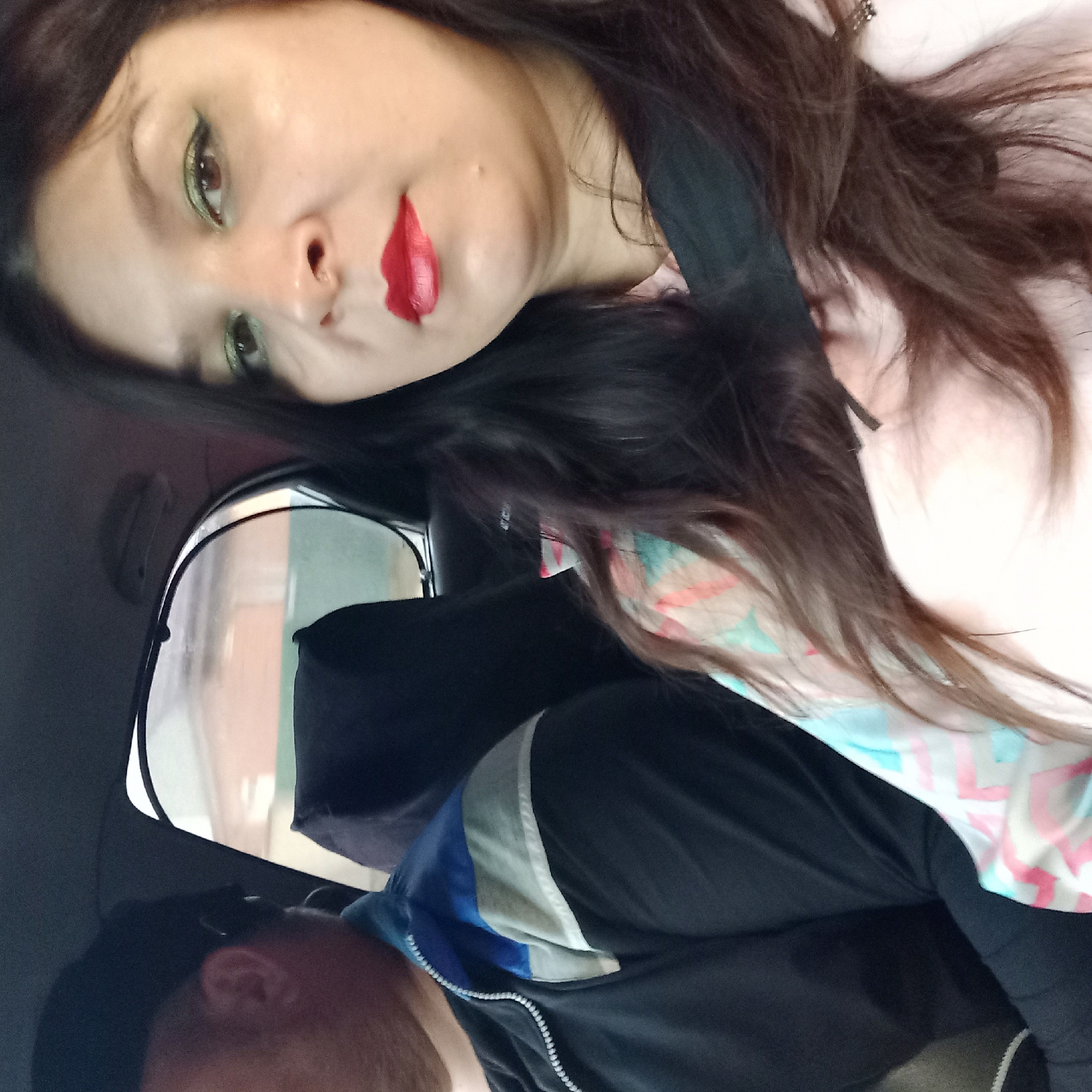 The image shows two people in the back seat of a vehicle, likely a car, with a window on the right side providing a view of a blurred, outdoor scene. The person on the left is wearing a black baseball cap and a dark jacket with blue and gray accents on the shoulder. The person on the right, a woman, is closer to the camera and facing it. She has long, dark hair, green eyeshadow, and red lipstick. She is wearing a light pink top with a patterned section on her shoulder that has colors like pink, blue, and white. Both individuals appear to be passengers, and the setting suggests they are traveling within the city of Kropyvnytskyi, Ukraine, based on the information provided. The overall lighting is natural, possibly from the daytime, and the interior of the car is dimly lit.