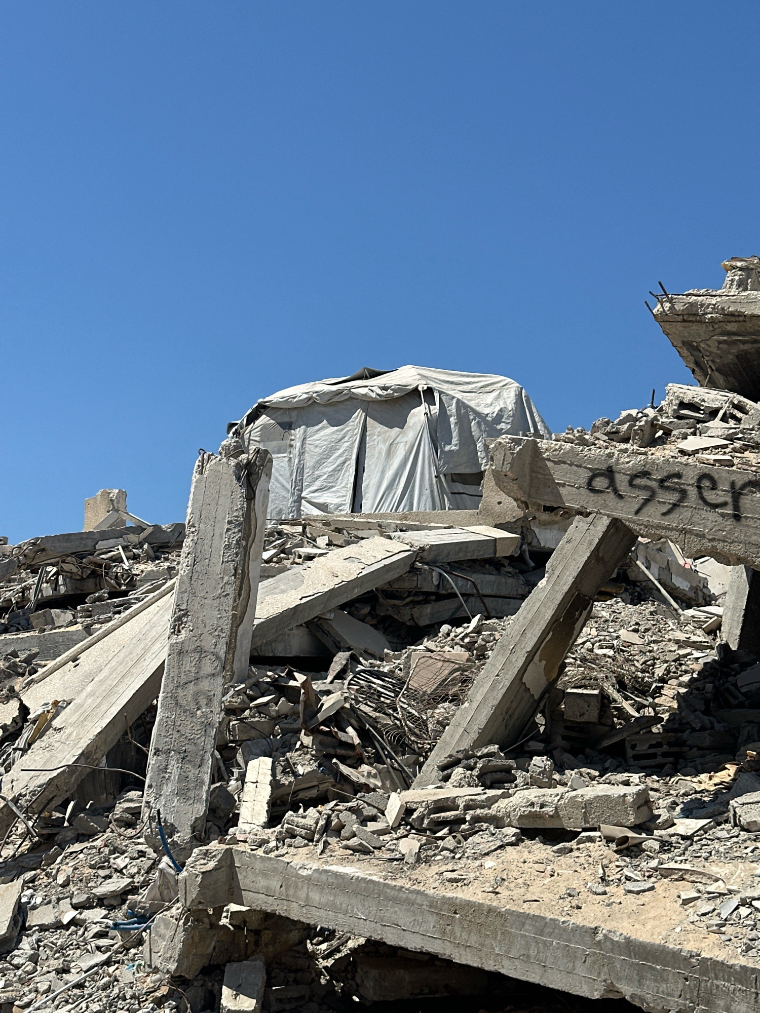 The image depicts a scene of destruction in the city of Gaza, Palestinian Territories. A white tent is set up amidst the rubble of what appears to be a collapsed building. The tent is partially covered by debris. The surrounding area is filled with broken concrete, rebar, and other building materials, indicating significant damage. In the background, the sky is clear and blue. There is black graffiti on a piece of concrete that says "asser". The overall scene conveys a sense of devastation and the precariousness of the current living conditions.