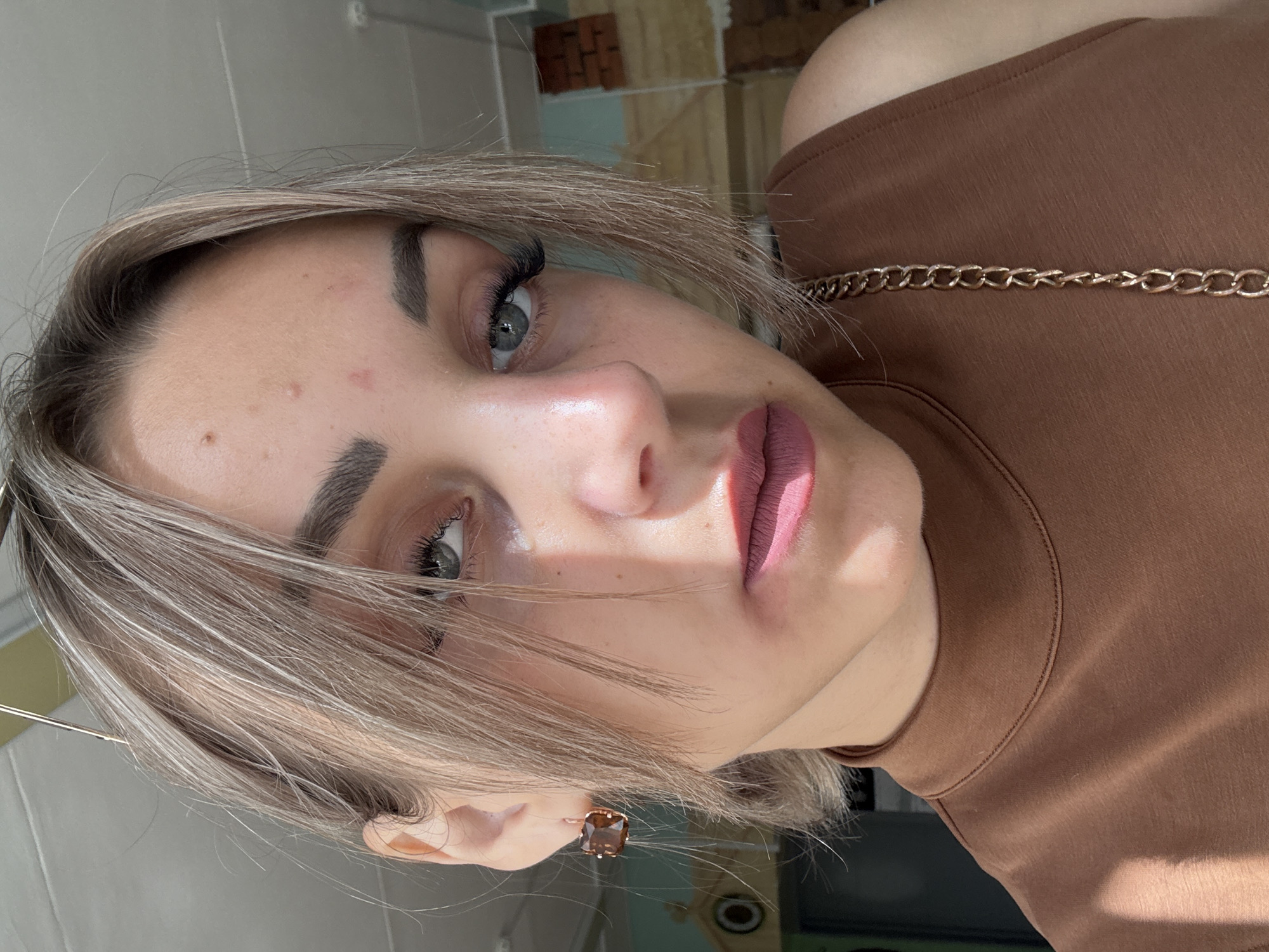 The image is a close-up, selfie-style portrait of a young woman with blonde hair and light blue eyes. She is wearing a brown sleeveless turtleneck top with a gold chain strap. Her makeup includes dark eyebrows, long eyelashes, and matte pink lipstick. She is also wearing a square brown earring. The background appears to be an indoor setting with a brick wall and a wooden surface. Sunlight is casting shadows on the woman and her clothing. There are no obvious elements in the image that indicate the specific location within the city of Yuzhno-Sakhalinsk, Russia.