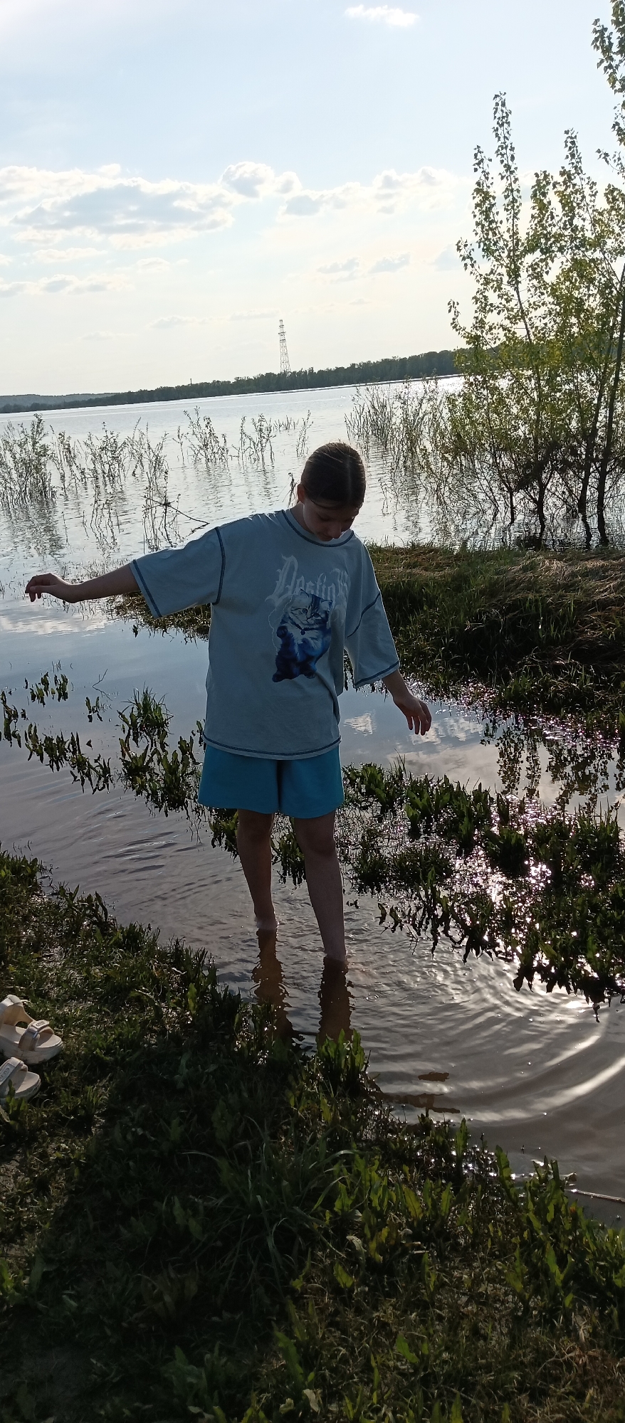 A person stands in shallow water at the edge of a lake or flooded area. The person is wearing a light blue t-shirt with a graphic on the front and matching blue shorts. Their feet are submerged in the water, and they have their arms extended to the sides. Vegetation grows in the water and along the shoreline. In the background, there is a body of water with trees and a distant utility pole visible on the horizon. A pair of sandals sit on the grass next to the water. The lighting indicates it is either early morning or late afternoon. The location is Unknown, Unknown.