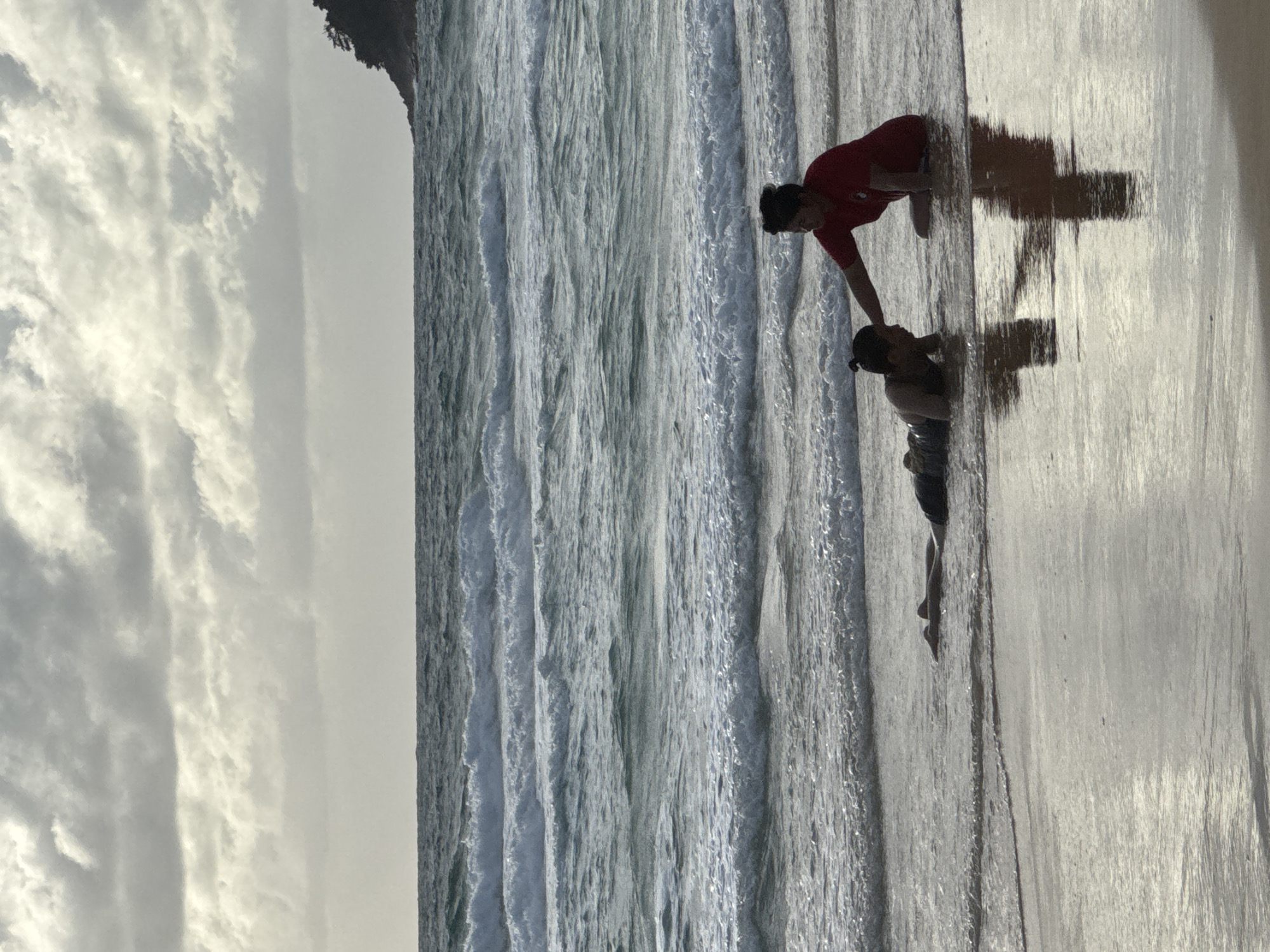 In Karon, Thailand, on a sandy beach, two individuals are present near the waterline. One person, positioned on their stomach in the shallow water, is wearing dark clothing. The other person, seated on their knees, is wearing a red shirt. The seated individual is reaching out towards the person on the ground, with their hand touching the other person's head. The ocean exhibits moderate waves, and the sky is overcast with clouds. The sand reflects the light, creating a shimmering effect.